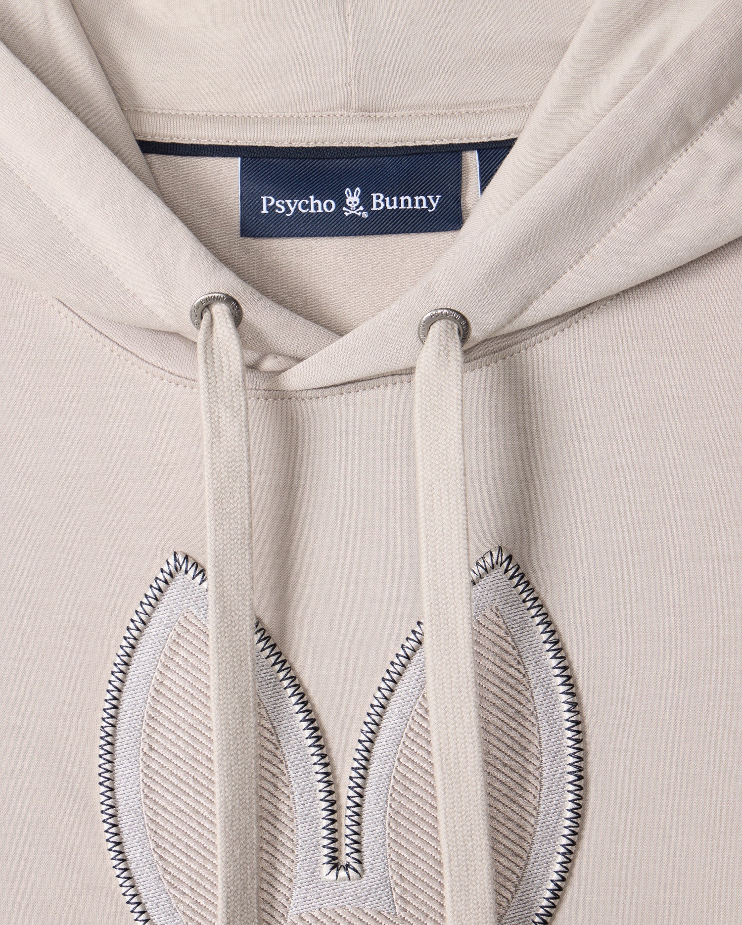 Close-up of the MENS WALTER LIGHTWEIGHT HOODIE - B6H476D200 by Psycho Bunny, crafted from beige Micro French terry. This regular fit hoodie features two drawstrings and an embroidered bunny ear design on the chest area, with the "Psycho Bunny" label sewn inside.