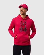 A person wearing the Psycho Bunny MENS WALTER LIGHTWEIGHT HOODIE - B6H476D200 in red, adorned with a bunny skull logo and intricate 3D outlined embroidery, stands with hands in the front pocket. They pair it with a matching red cap and black pants, smiling against a plain background.