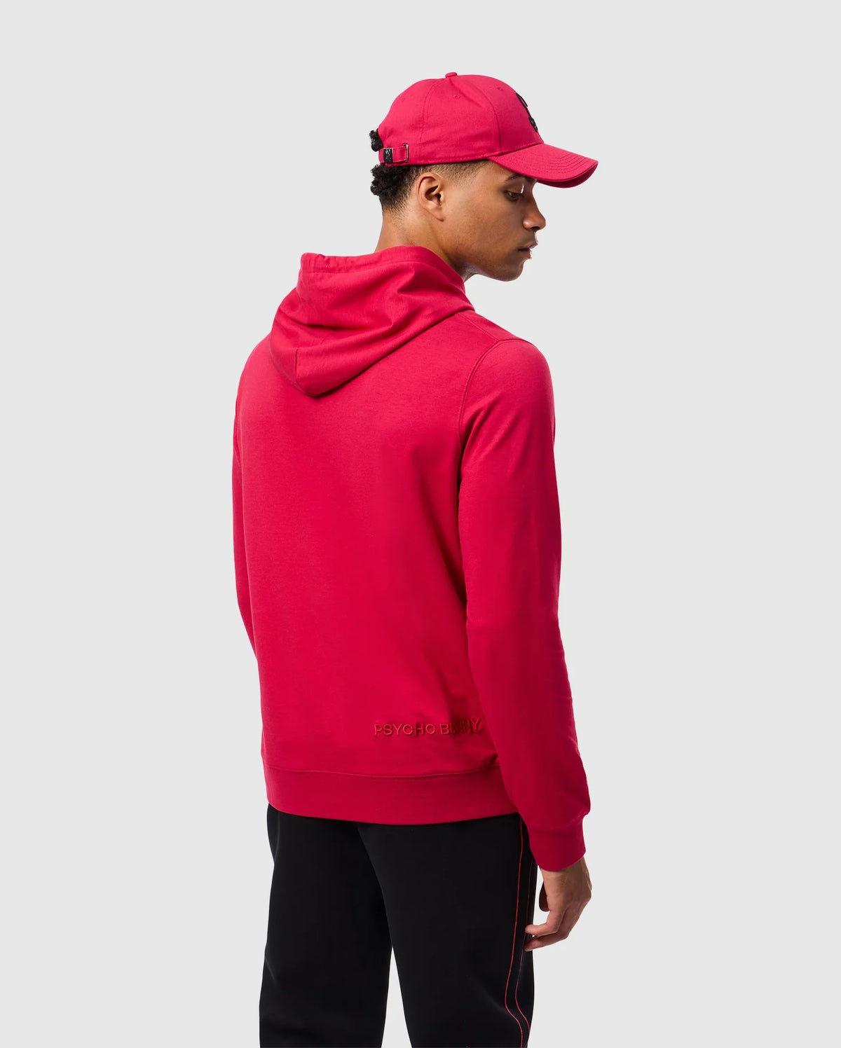 A person wearing a Psycho Bunny MENS WALTER LIGHTWEIGHT HOODIE - B6H476D200 in red with 3D outlined embroidery and a matching cap, paired with black pants, stands with their back turned and gazes slightly to the side against a plain gray background.