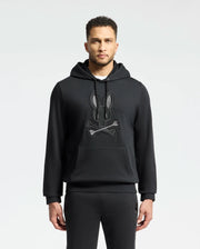 A man in an elegant Psycho Bunny MENS NEW CASTLE FRENCH TERRY HOODIE - B6H517F200, showcasing a stylized rabbit and bones design on the front, stands against a plain light background.