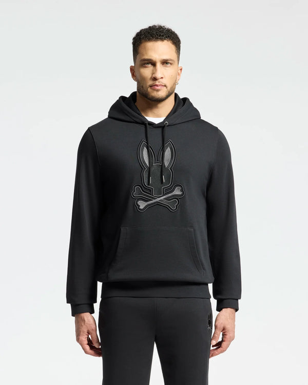 A man in an elegant Psycho Bunny MENS NEW CASTLE FRENCH TERRY HOODIE - B6H517F200, showcasing a stylized rabbit and bones design on the front, stands against a plain light background.