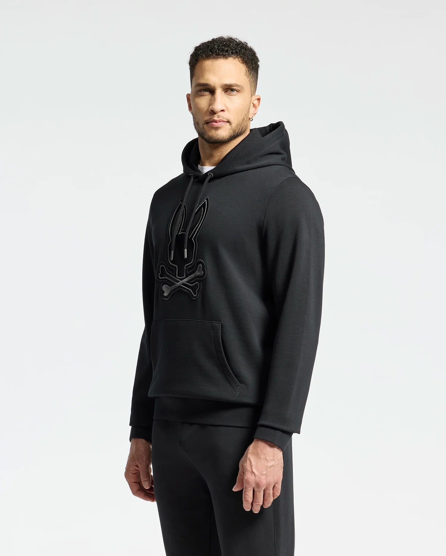 A man in a Psycho Bunny MENS NEW CASTLE FRENCH TERRY HOODIE (B6H517F200) with a rabbit skull and crossbones design stands against a plain light background, looking at the camera. Crafted from cozy French terry, it combines comfort with edgy sophistication.
