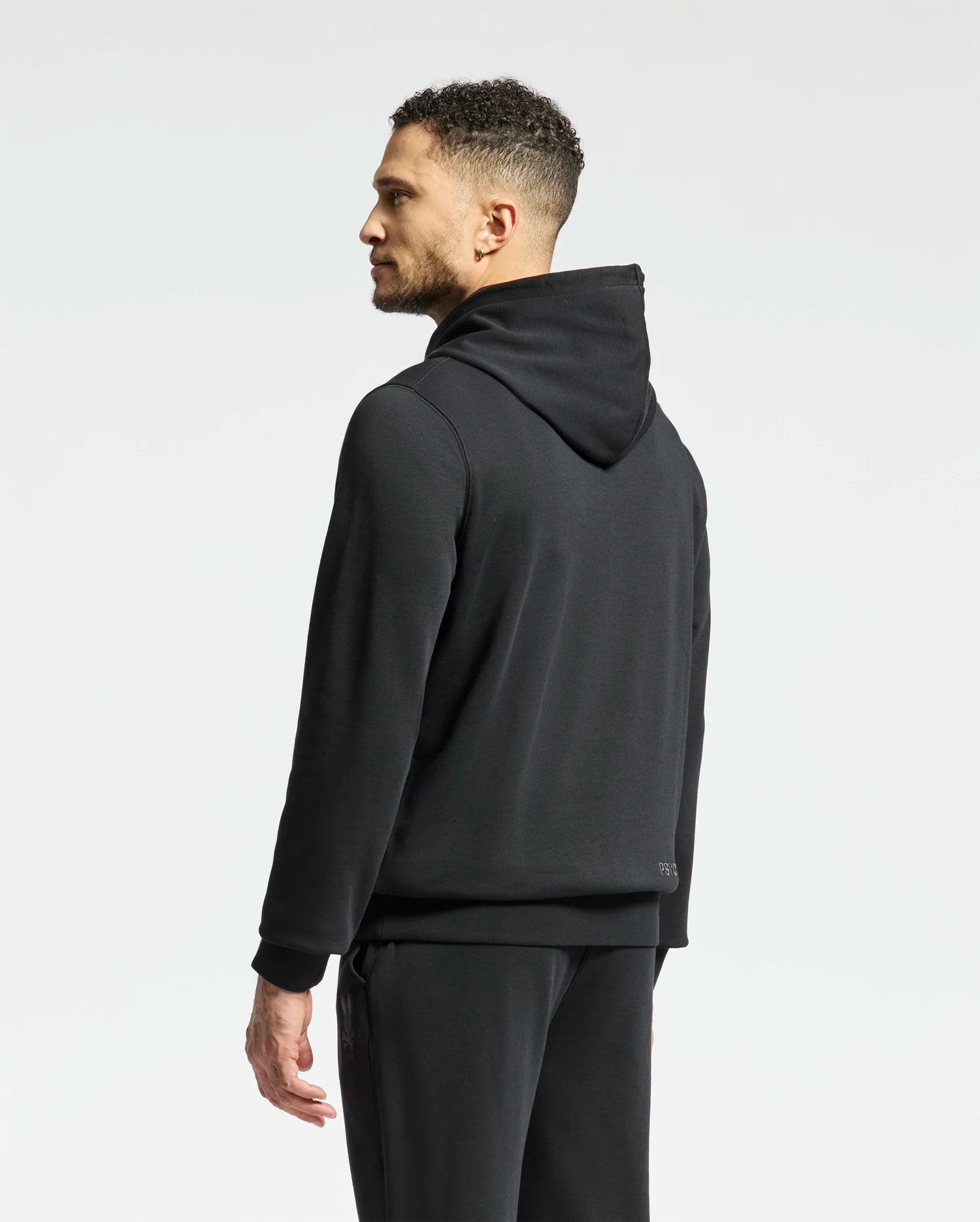 Against a plain gray background, a person with short curly hair showcases an elegant style in a black Psycho Bunny MENS NEW CASTLE FRENCH TERRY HOODIE and matching pants, hood up. The overall look is casual yet athletic.