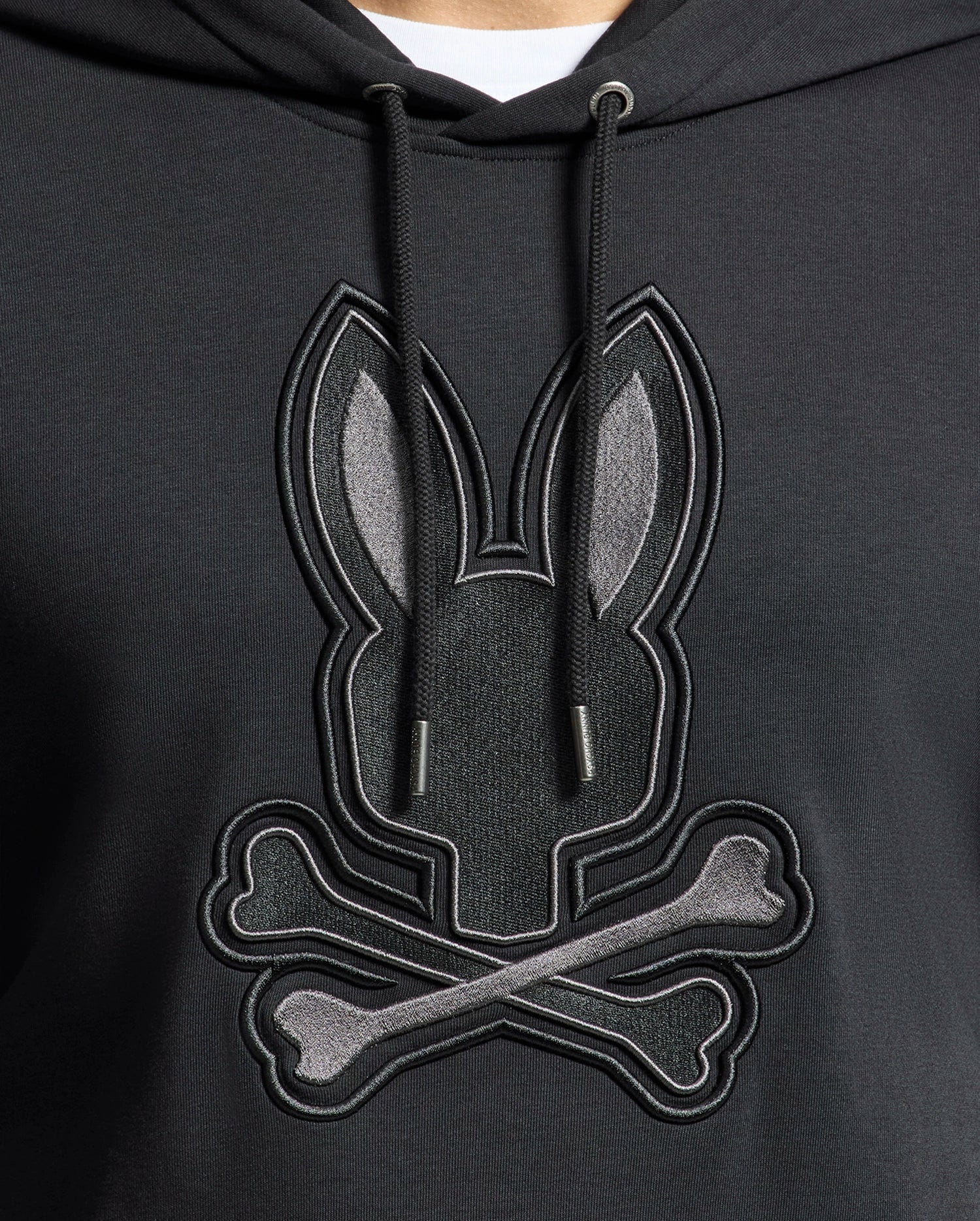 A close-up of Psycho Bunny's MENS NEW CASTLE FRENCH TERRY HOODIE (B6H517F200): an elegant black hoodie with a soft French terry fabric, showcasing a stylized embroidered bunny head and crossbones motif in gray shades on the front.