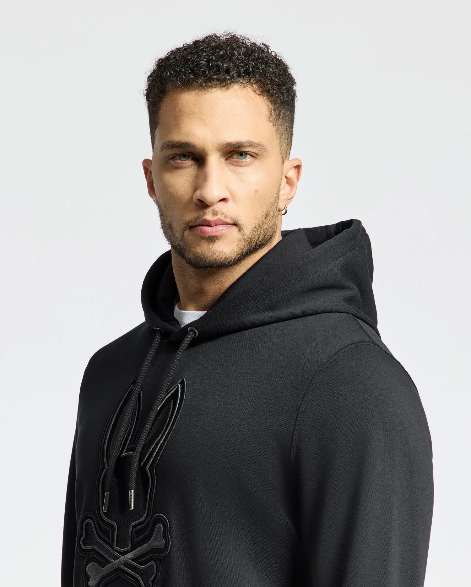 A man wearing a Psycho Bunny Men's New Castle French Terry Hoodie (B6H517F200) in black with a rabbit skull and crossbones design, gazes directly at the camera. He has short curly hair and an earring, set against a plain white background.
