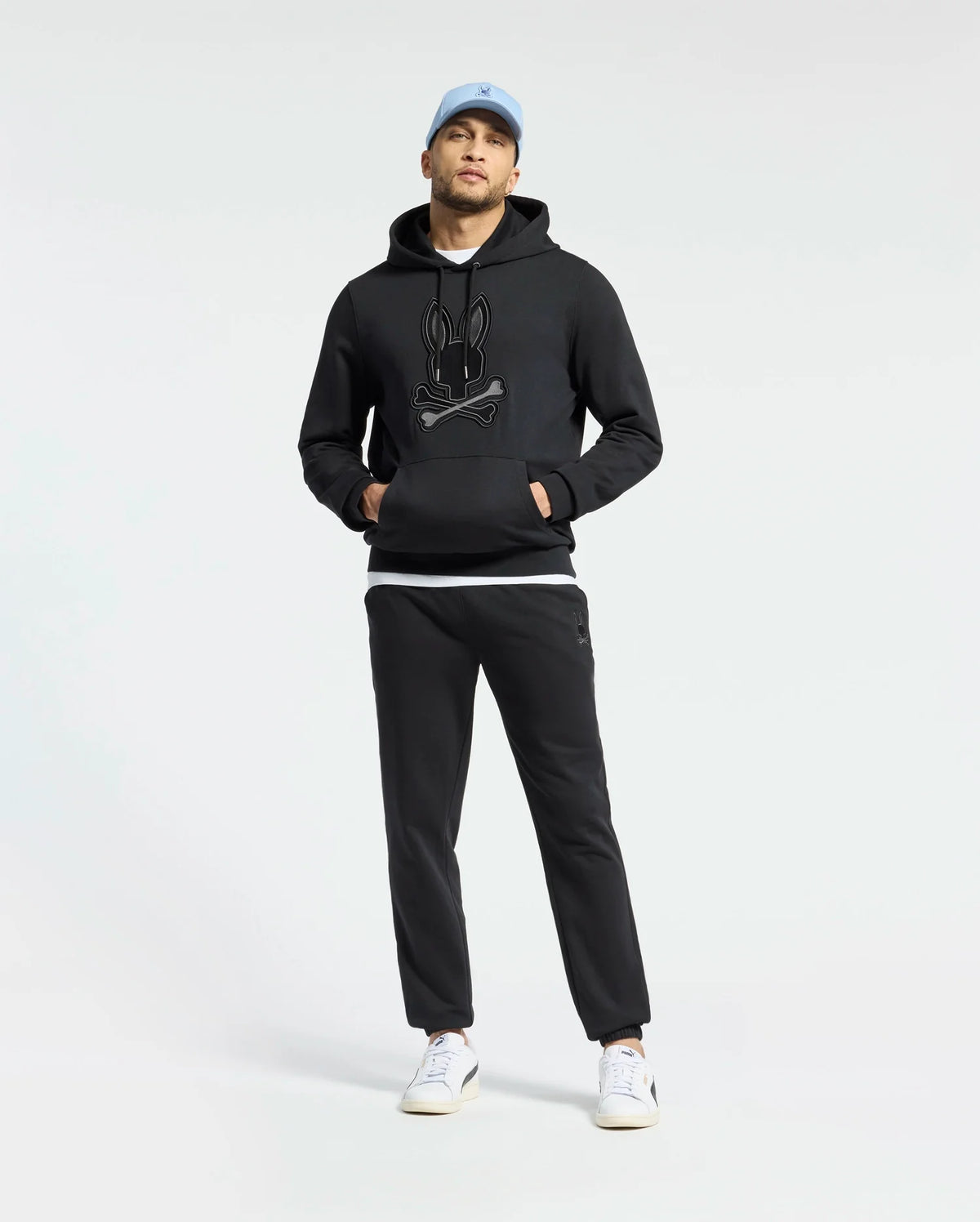 A person in a black Psycho Bunny Men's New Castle French Terry Hoodie pairs it with black pants and white sneakers, completing the look with a light blue cap against a plain white background.