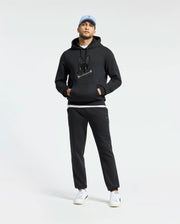 A person in a black Psycho Bunny Men's New Castle French Terry Hoodie pairs it with black pants and white sneakers, completing the look with a light blue cap against a plain white background.