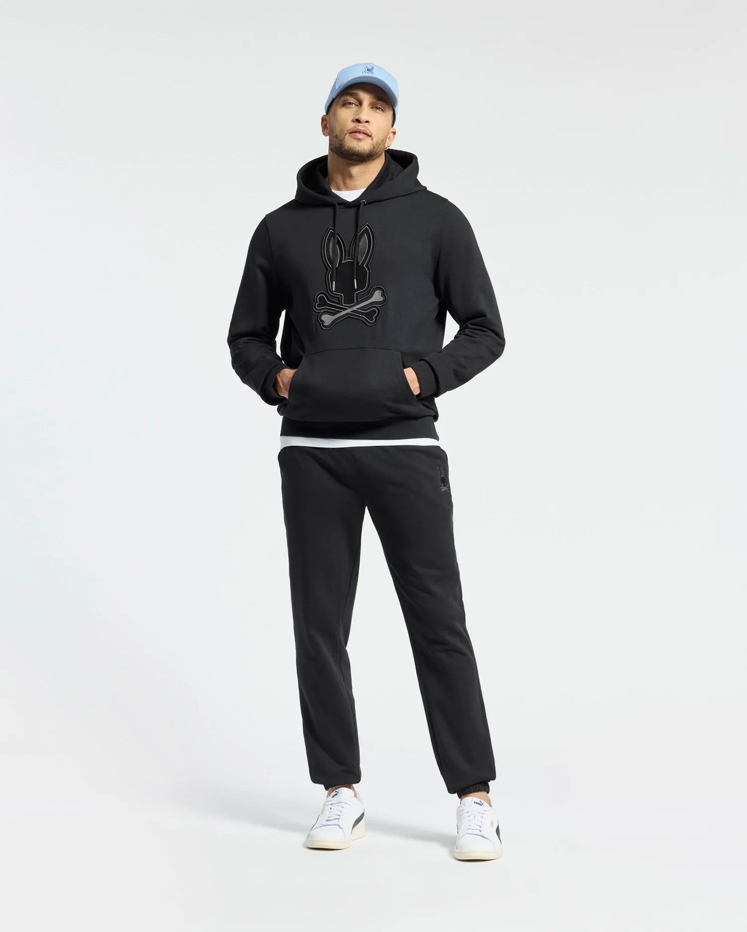 A person in a black Psycho Bunny Men's New Castle French Terry Hoodie pairs it with black pants and white sneakers, completing the look with a light blue cap against a plain white background.