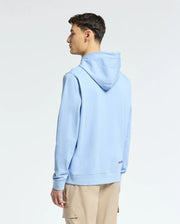 A person stands slightly left, wearing a Psycho Bunny's Mens New Castle Embroidered French Terry Hoodie (B6H517F200) in light blue with beige pants. The hood is down against a plain white backdrop, showing the side and back view.