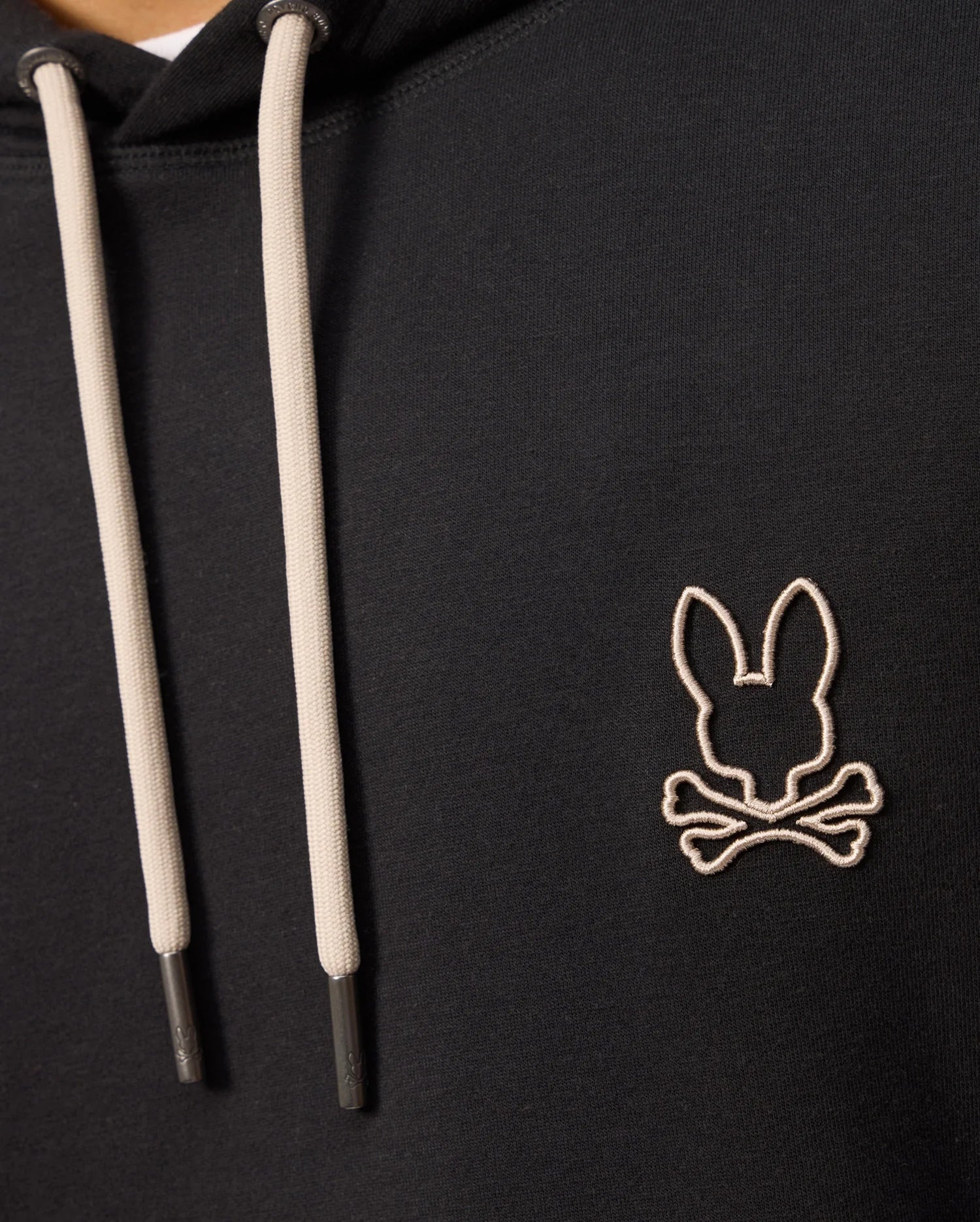 Close-up of a Psycho Bunny MENS JAMES PREMIUM HOODIE - B6H789D200 in black, featuring cream-colored drawstrings. The hoodie showcases an embroidered bunny logo on the left chest area, depicting a stylized rabbit head above crossed bones, outlined in white stitching.