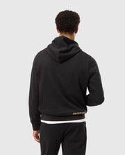 A person with short curly hair is facing away from the camera, wearing a Psycho Bunny MENS JAMES PREMIUM HOODIE - B6H789D200 in black along with black pants. The hoodie features the text "PSYCHO BUNNY" in small letters near the waistline and an embroidered bunny on the back. The background is plain and light-colored.