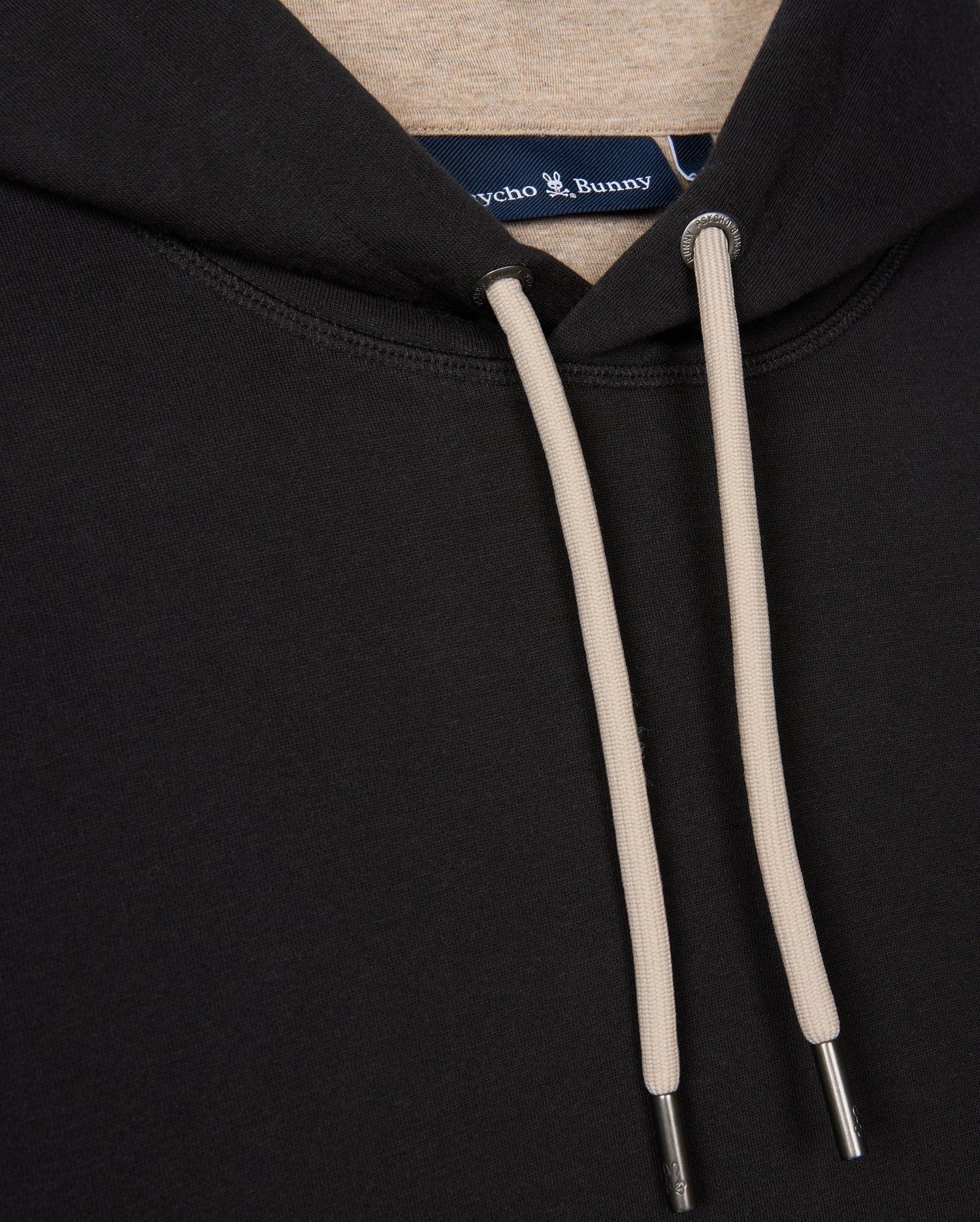 Close-up of the neck area of the MENS JAMES PREMIUM HOODIE - B6H789D200 by Psycho Bunny, featuring black fabric with beige drawstrings. The focus is on the hoodie and its intricate details, including precise stitching and metal tips on the drawstrings. A portion of the interior label with an embroidered bunny is also visible.