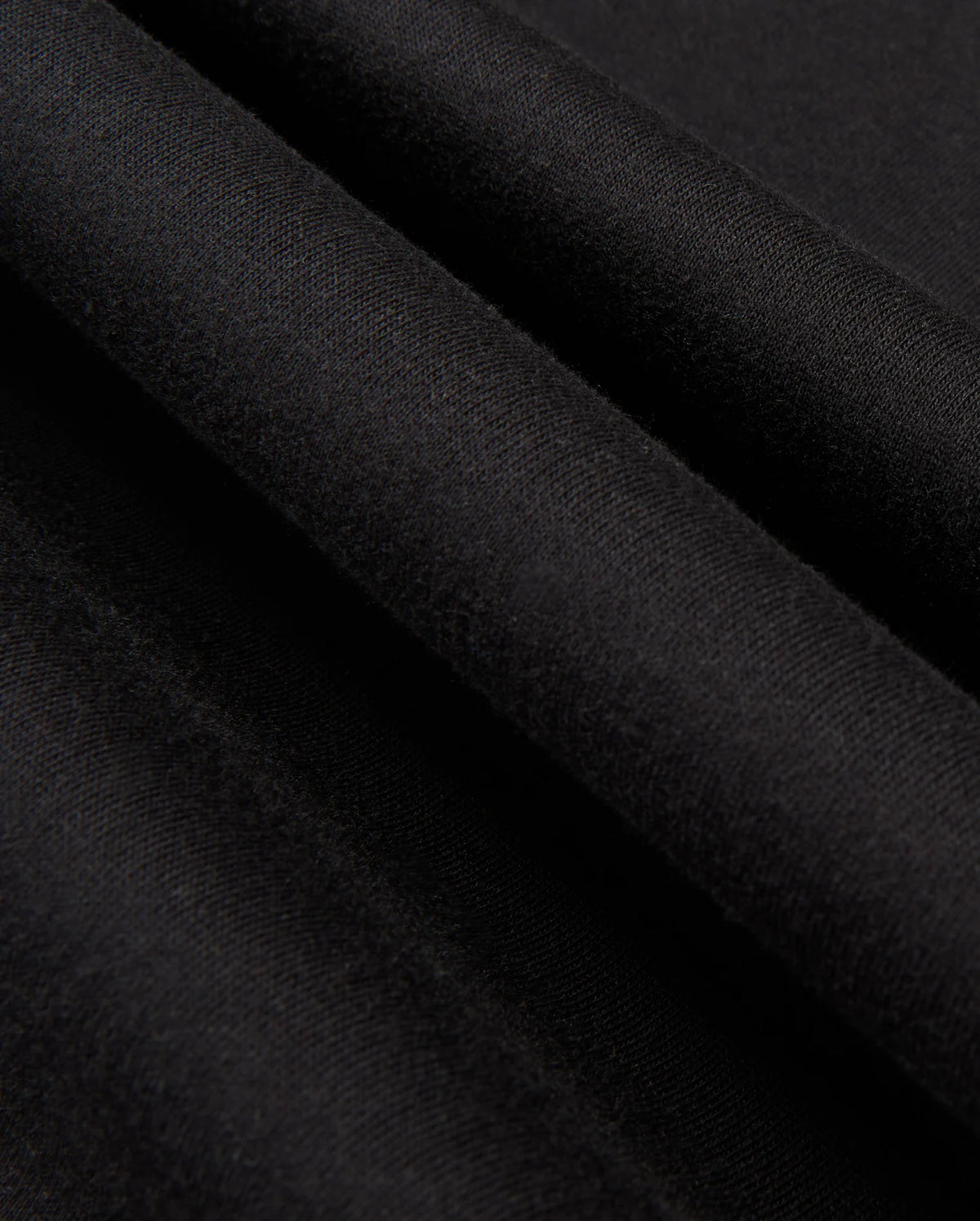 A close-up view of the smooth, black fabric from the Psycho Bunny MENS JAMES PREMIUM HOODIE - B6H789D200 reveals three parallel folds running diagonally across the image. The texture appears soft and even, resembling essential fleece, with a matte finish that creates a subtle interplay of light and shadow on the surface.