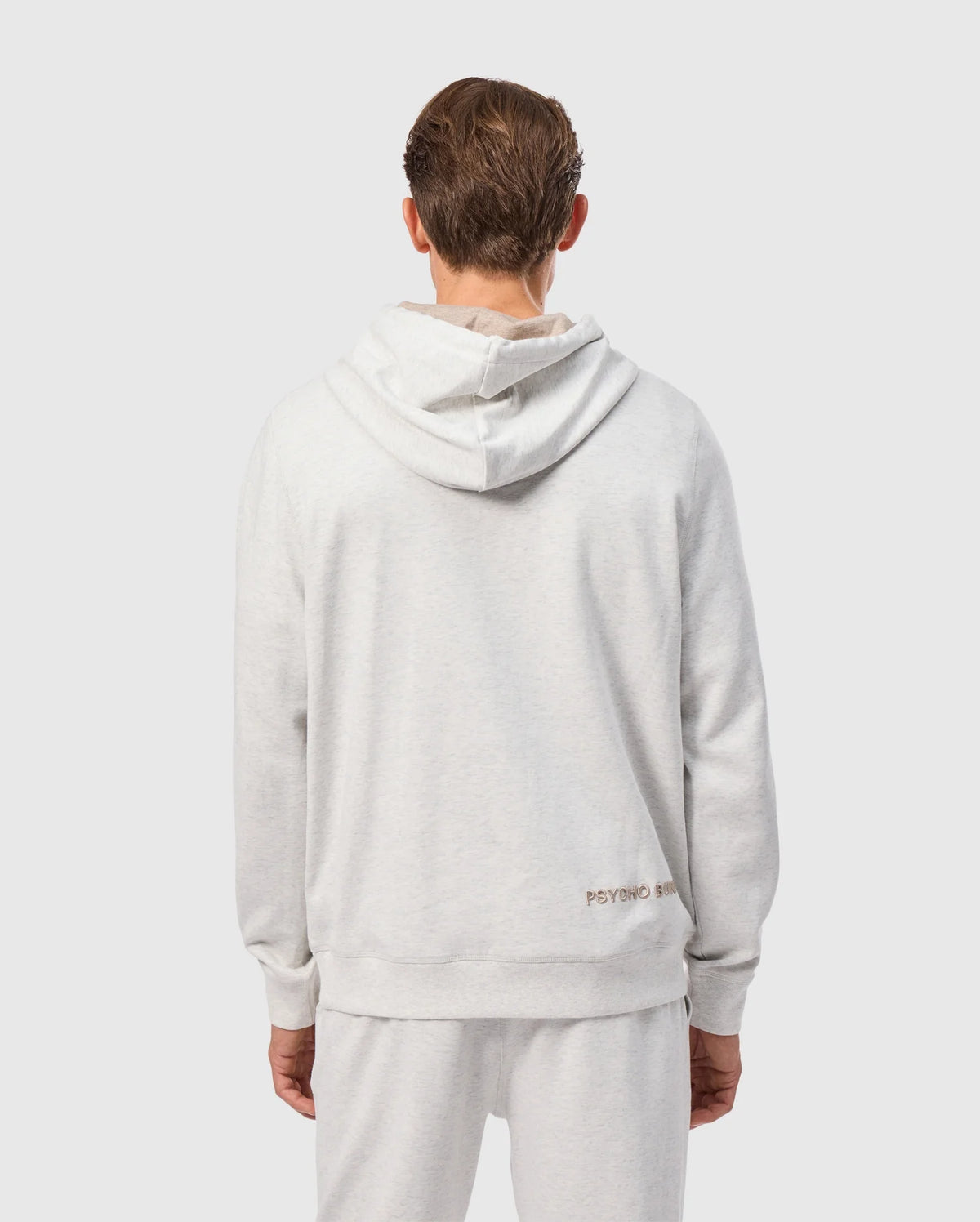 A person is shown from the back wearing the MENS JAMES PREMIUM HOODIE - B6H789D200 by Psycho Bunny in light gray, paired with matching pants. This premium jersey hoodie is adorned with an embroidered bunny near the bottom, adding a touch of stylish warmth. The background is plain and light-colored.