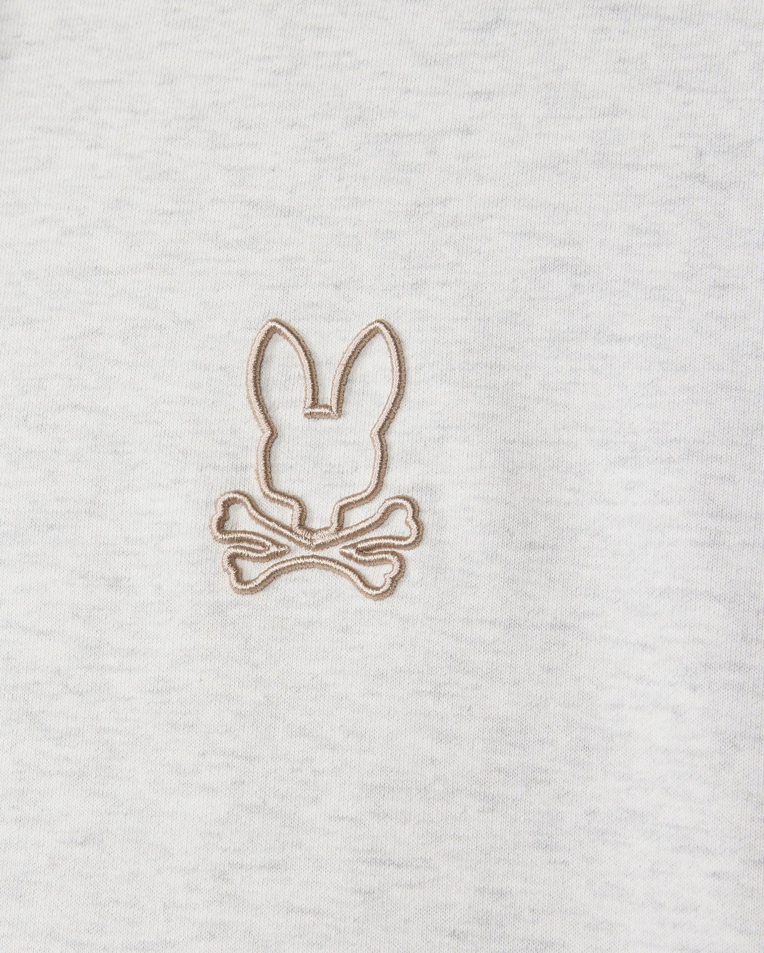 Highlighted in a detailed close-up, the MENS JAMES PREMIUM HOODIE (B6H789D200) from Psycho Bunny features an embroidered bunny design with a rabbit's head outline above two crossed bones. Beige thread contrasts subtly against the gray jersey fabric, infusing stylish warmth into this premium piece.