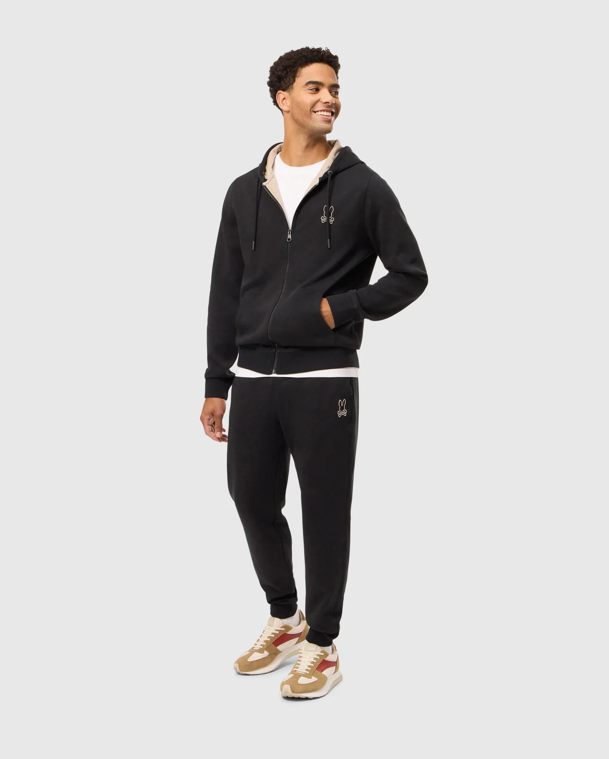 A man wearing a Psycho Bunny MENS JAMES PREMIUM ZIP HOODIE - B6H793D200 in double-faced premium jersey, a white undershirt, and black joggers stands looking to his left with his right hand in the hoodie pocket. He is also sporting beige and red sneakers against a plain white background.