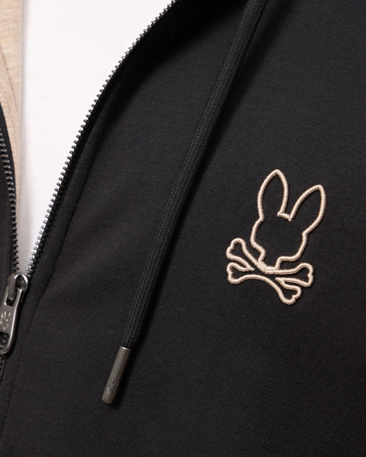 Close-up of the Psycho Bunny MENS JAMES PREMIUM ZIP HOODIE - B6H793D200, showcasing an embroidered Bunny graphic over crossed bones on the right side of the chest. Made from double-faced premium jersey, it features a zipper on the left side and a drawstring with metal tips.
