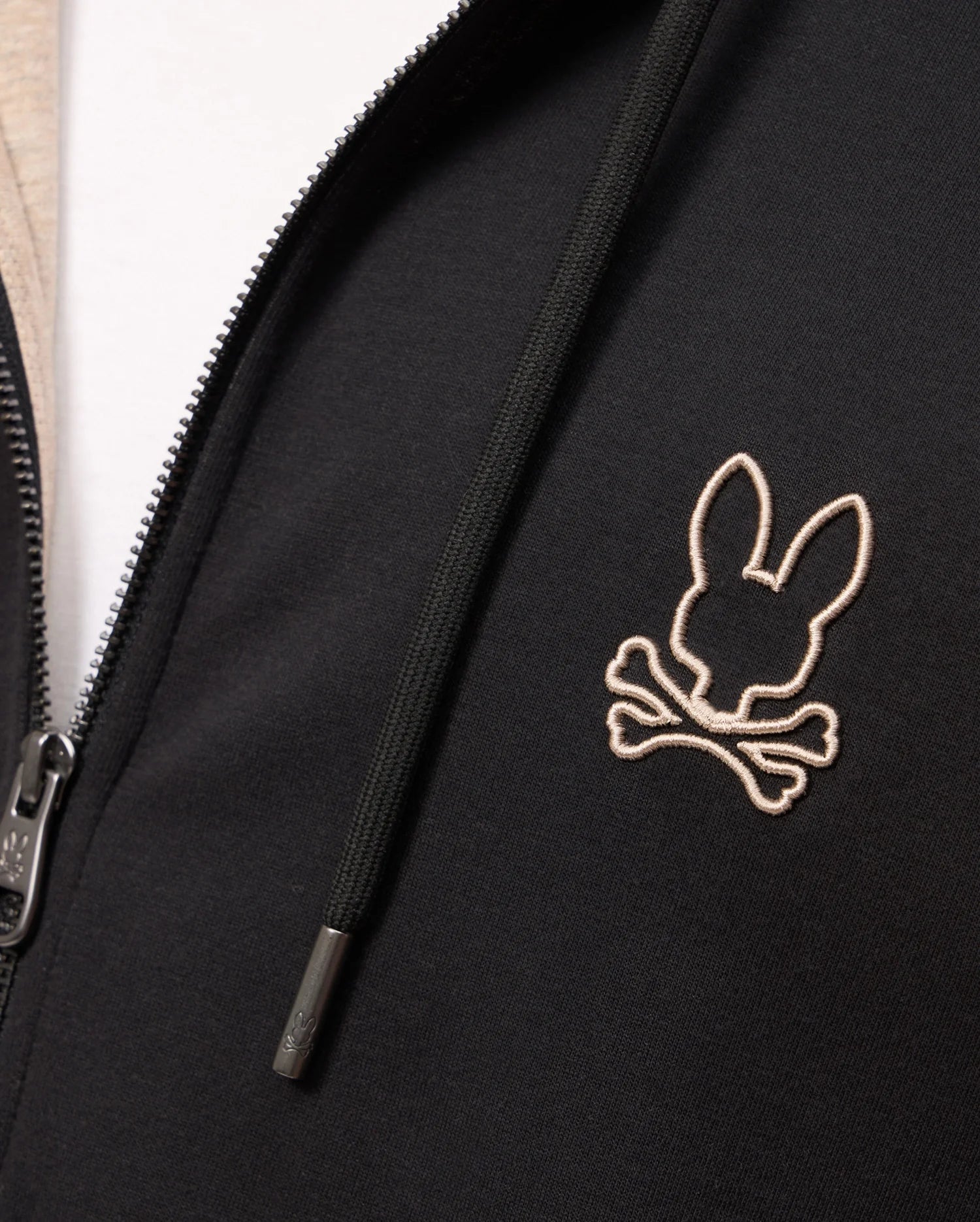 Close-up of the Psycho Bunny MENS JAMES PREMIUM ZIP HOODIE - B6H793D200, showcasing an embroidered Bunny graphic over crossed bones on the right side of the chest. Made from double-faced premium jersey, it features a zipper on the left side and a drawstring with metal tips.