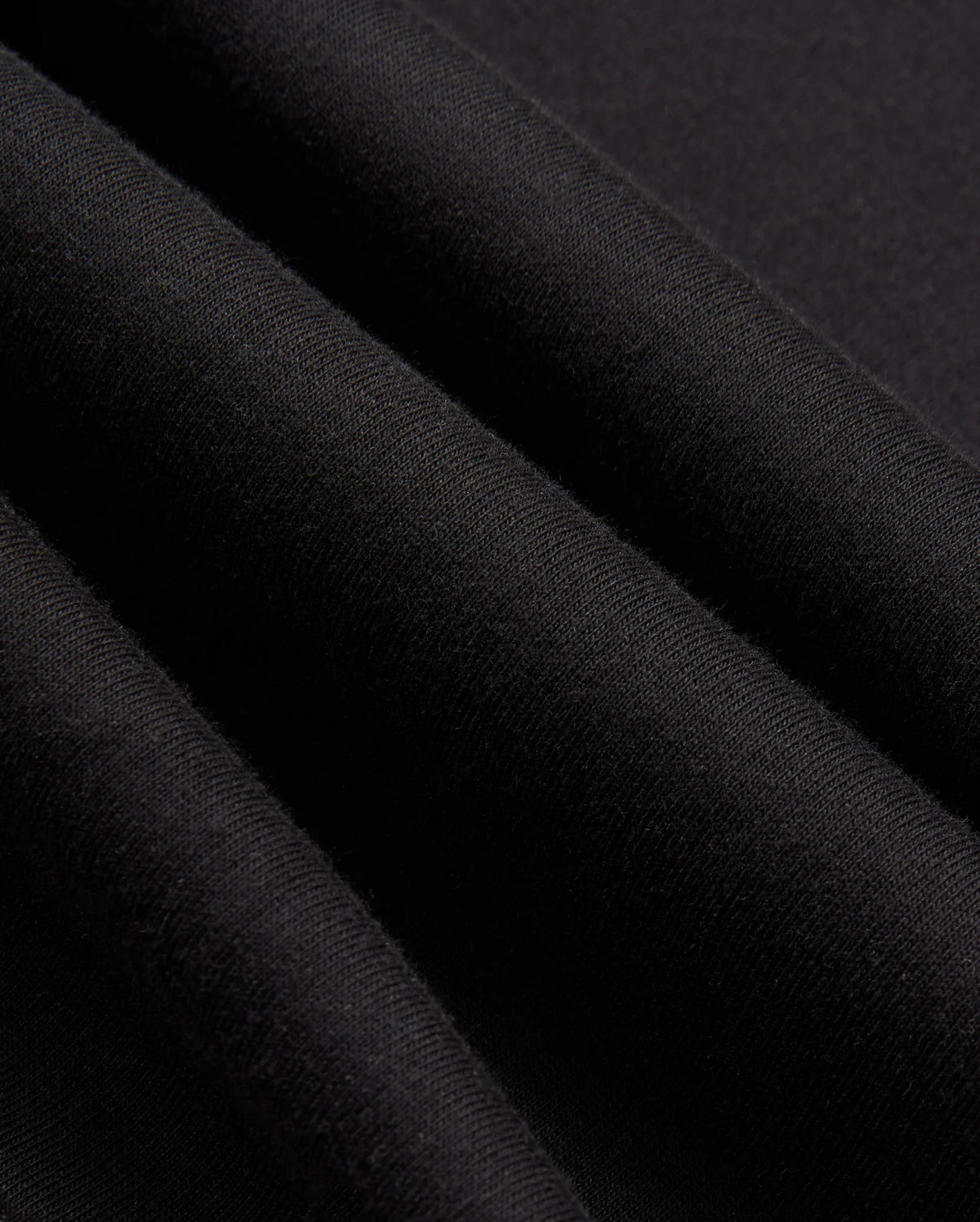 Close-up of the black fabric from the MENS JAMES PREMIUM ZIP HOODIE - B6H793D200 by Psycho Bunny showing its texture. The fabric appears soft and slightly folded, creating gentle shadows and highlights. The material, part of a double-faced premium jersey, looks smooth and uniform in color.