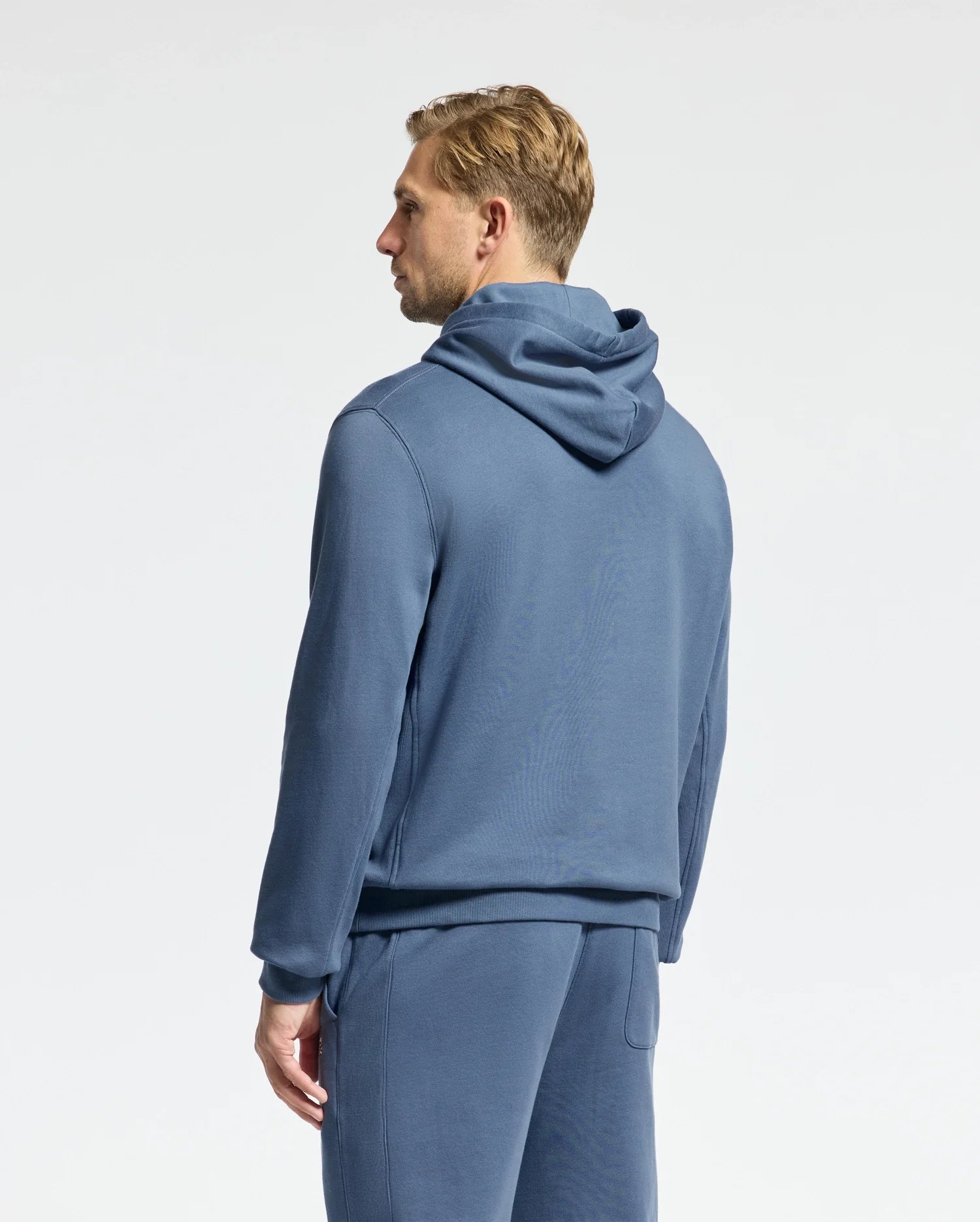 A person wearing a blue Psycho Bunny MENS LEVY ESSENTIAL FLEECE HOODIE - B6H859D200 and matching pants stands facing away against a plain background, showcasing a side profile. Crafted from 100% cotton French terry, the ensemble complements the individual's short, light-colored hair.