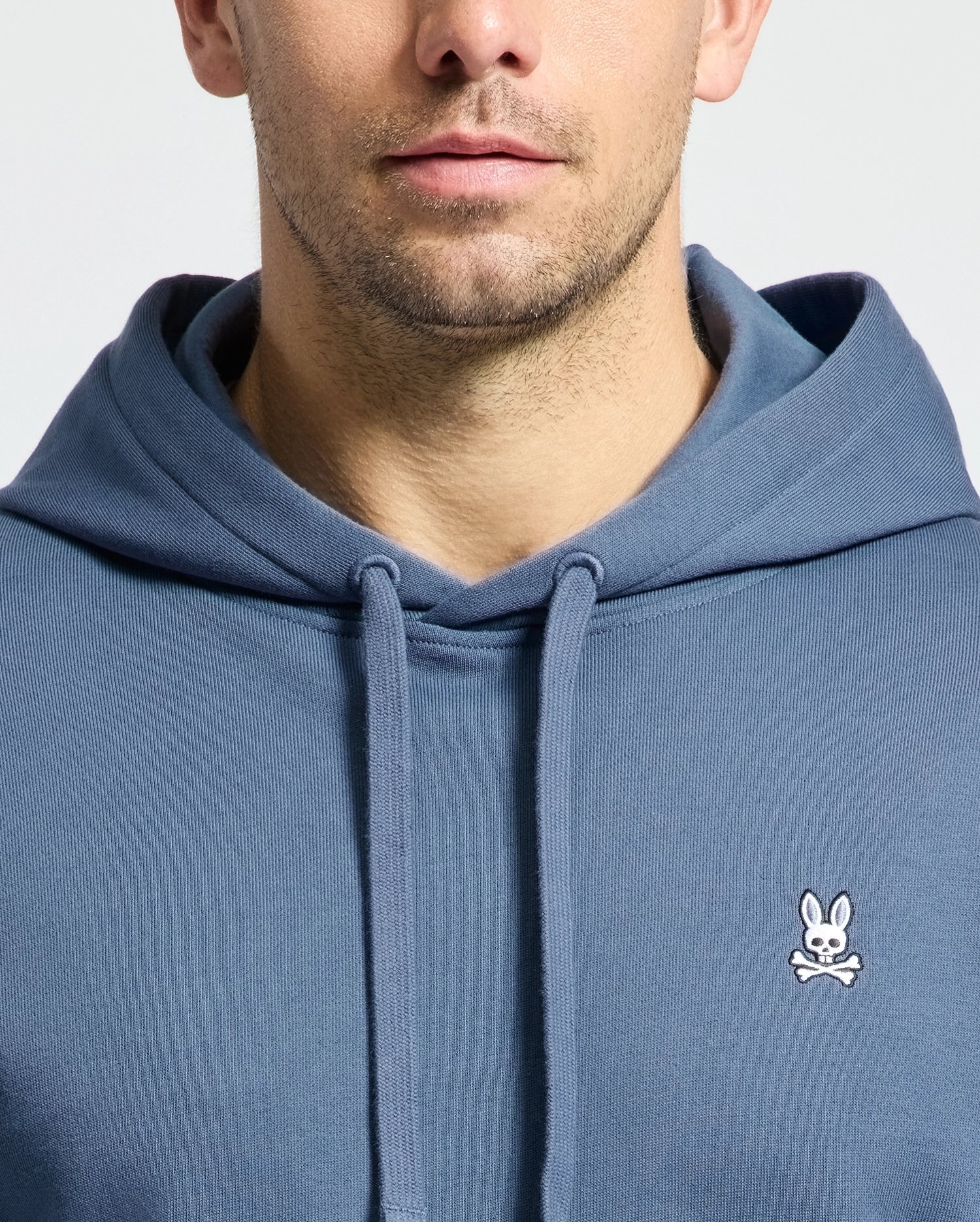 A man is seen wearing the blue MENS LEVY ESSENTIAL FLEECE HOODIE - B6H859D200 by Psycho Bunny, constructed from soft French terry and adorned with a small white bunny and crossbones logo on the chest. The image shows the lower half of his face and upper chest against a simple background.