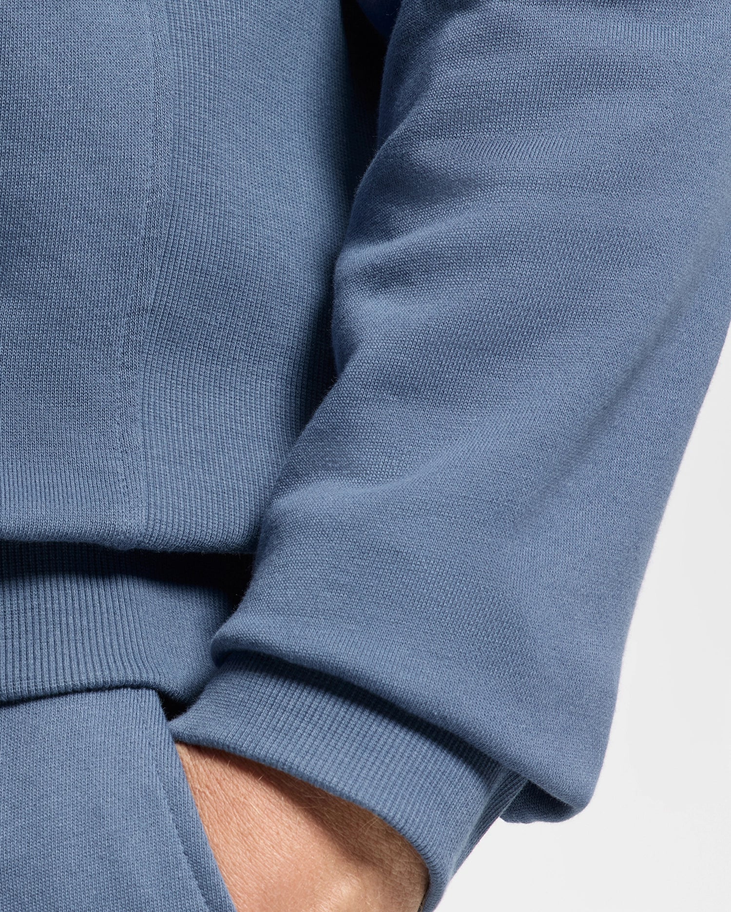 Close-up of a person wearing the MENS LEVY ESSENTIAL FLEECE HOODIE by Psycho Bunny in blue, with their hand partially tucked into a matching pocket. The 100% cotton fabric looks ribbed and comfortable.