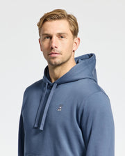 A person with short blond hair is wearing the MENS LEVY ESSENTIAL FLEECE HOODIE - B6H859D200 by Psycho Bunny. This blue men's hoodie is made from 100% cotton French terry and features a small bunny logo on the chest. They are looking directly at the camera against a plain light background.