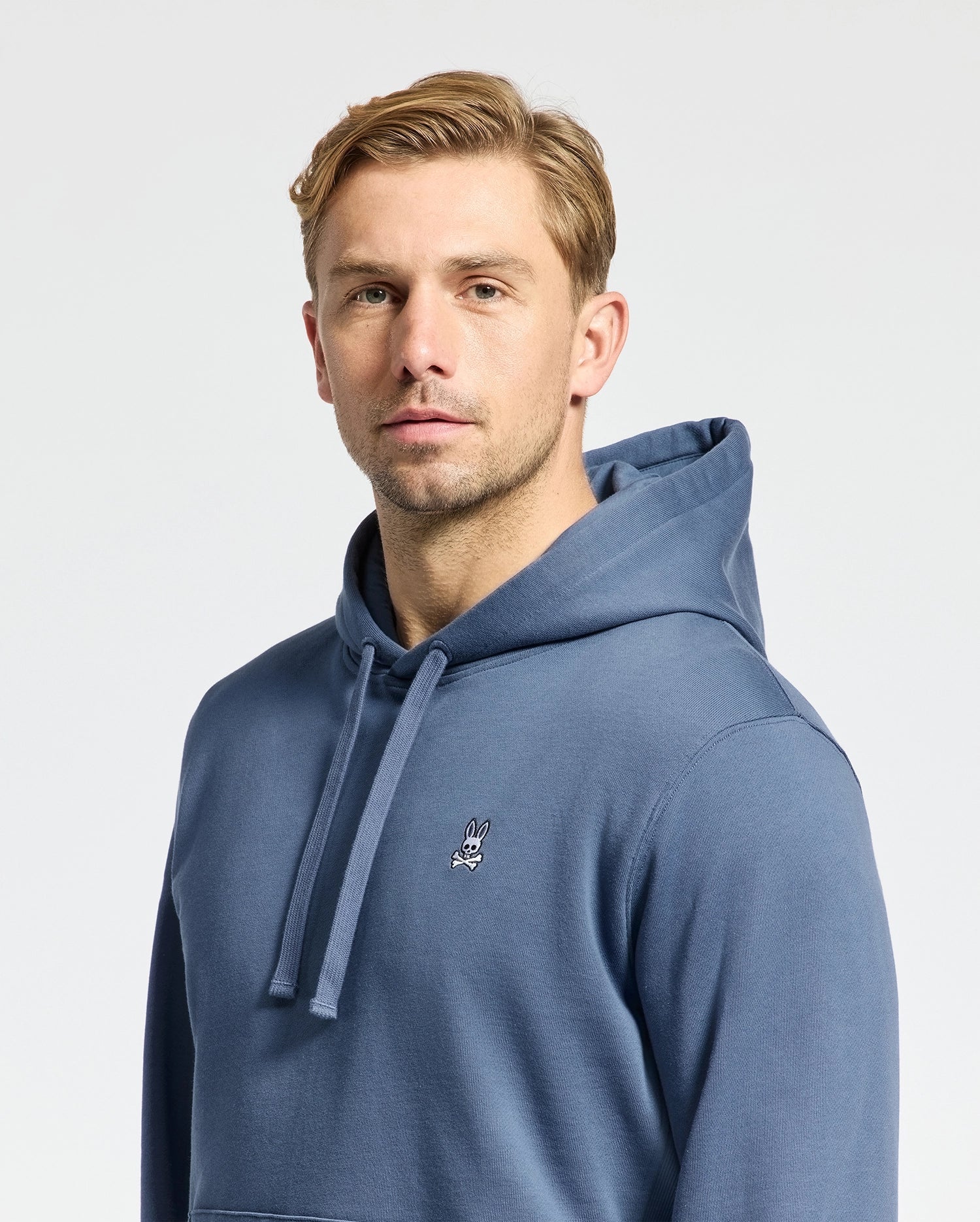 A person with short blond hair is wearing the MENS LEVY ESSENTIAL FLEECE HOODIE - B6H859D200 by Psycho Bunny. This blue men's hoodie is made from 100% cotton French terry and features a small bunny logo on the chest. They are looking directly at the camera against a plain light background.