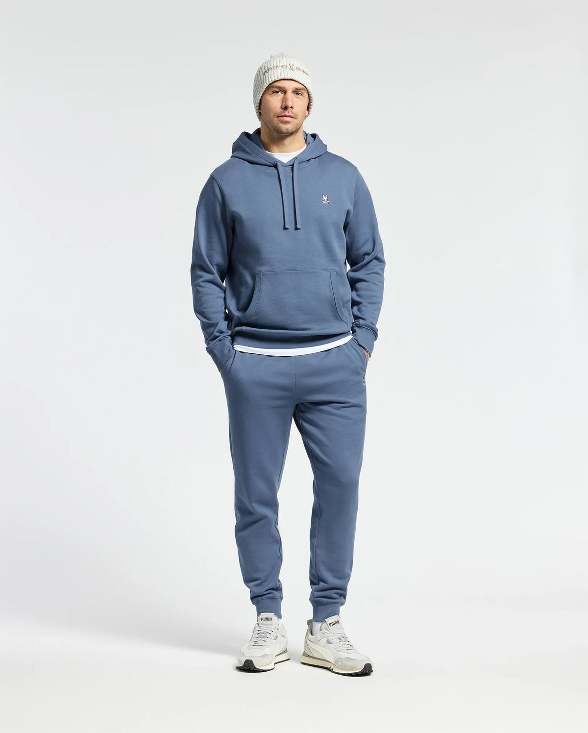 A man stands against a white background wearing the Psycho Bunny MENS LEVY ESSENTIAL FLEECE HOODIE - B6H859D200 in blue, paired with matching joggers, a white knit beanie, and 100% cotton white sneakers. His hands are in the hoodie pockets.