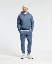 A man stands against a white background wearing the Psycho Bunny MENS LEVY ESSENTIAL FLEECE HOODIE - B6H859D200 in blue, paired with matching joggers, a white knit beanie, and 100% cotton white sneakers. His hands are in the hoodie pockets.