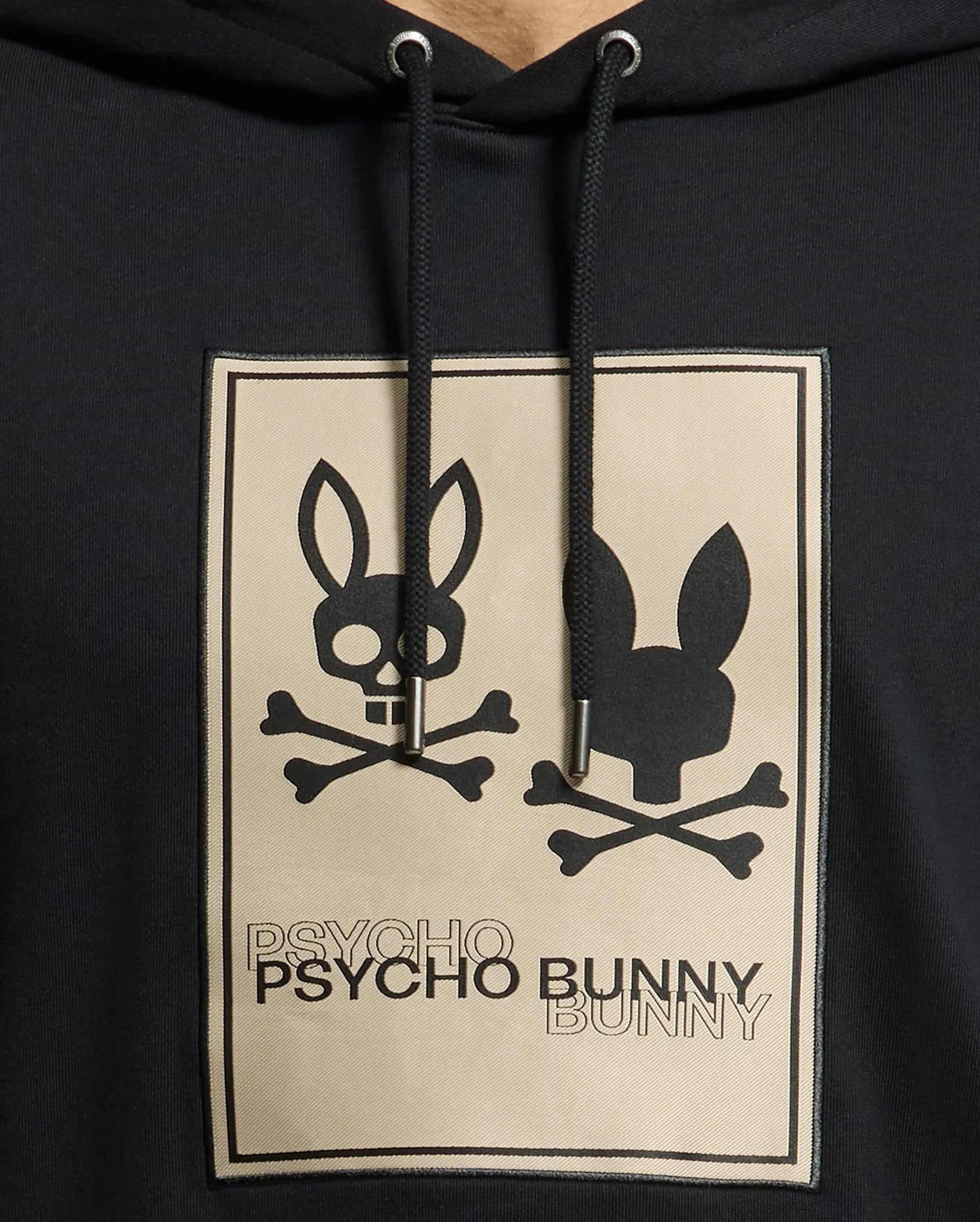Introducing the **MENS MONROE TWILL PATCH FLEECE HOODIE - B6H926D200** from *Psycho Bunny*. This stylish black hoodie is crafted from French terry fabric and features a unique graphic design of two bunny skulls. One skull showcases ear bones, while the other exhibits shaded ears over crossed bones. The "Psycho Bunny" text seamlessly integrates comfort with an edgy style statement.