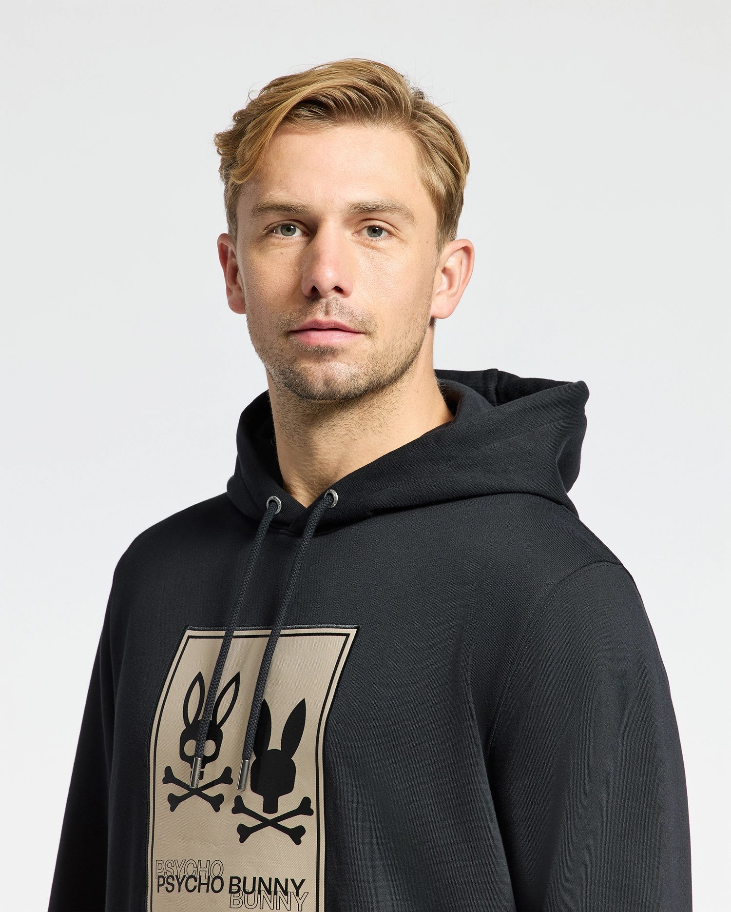 A man with short blonde hair models the edgy and comfortable Psycho Bunny MENS MONROE TWILL PATCH FLEECE HOODIE - B6H926D200, showcasing its unique skull and bunny graphic. He stands against a plain light background, gazing neutrally at the camera.