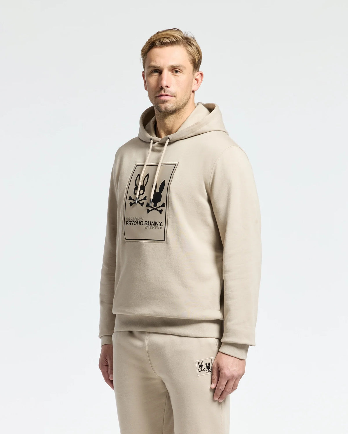 A man is wearing a Psycho Bunny MENS MONROE TWILL PATCH FLEECE HOODIE in beige, along with matching sweatpants, both featuring the brand's logo appliqué. He stands against a plain white background, looking at the camera with a neutral expression.