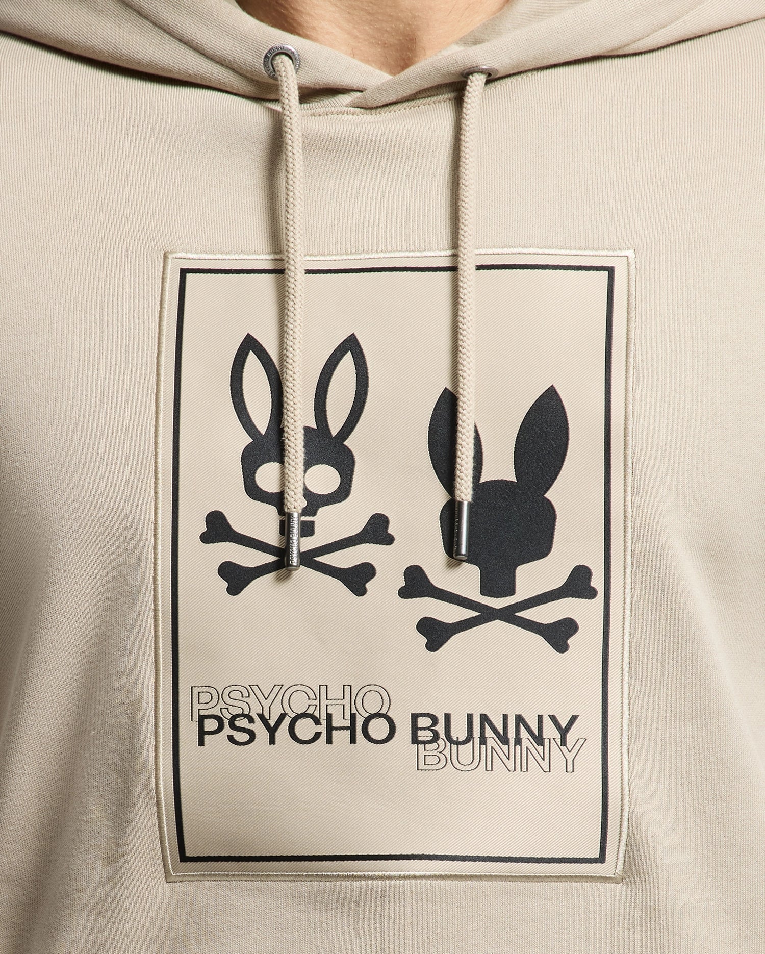 Close-up of the Psycho Bunny Men's Monroe Twill Patch Fleece Hoodie - B6H926D200 in beige, crafted from soft French terry. It features the iconic Psycho Bunny logo appliqué with two stylized bunny skulls and crossbones, along with the bold black text "PSYCHO" and "BUNNY" below the images.