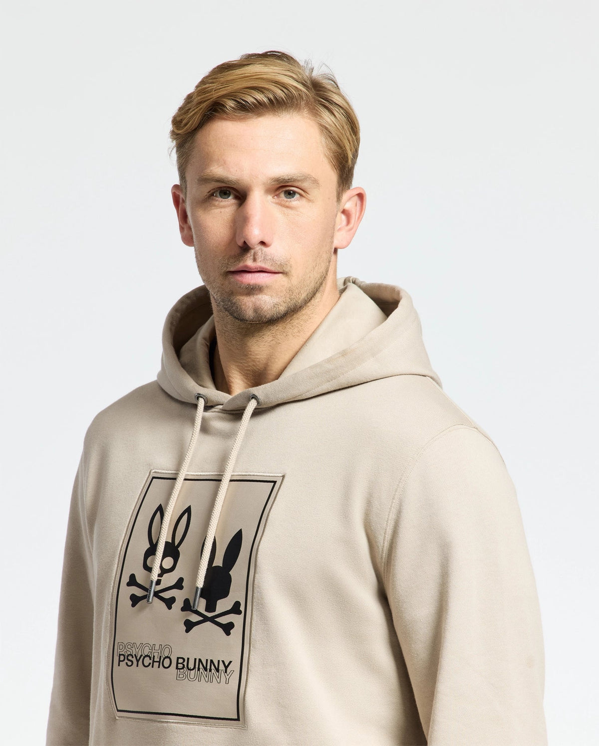 A man wearing a Psycho Bunny MENS MONROE TWILL PATCH FLEECE HOODIE - B6H926D200, adorned with a black graphic of a bunny and crossbones design, features the iconic "Psycho Bunny" logo appliqué, standing out against the plain white background.