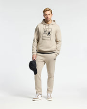 A man stands against a plain background, wearing a beige Psycho Bunny MENS MONROE TWILL PATCH FLEECE HOODIE and matching joggers. He holds a black cap in his right hand and wears white sneakers. The hoodie features a graphic design with logo appliqué on the front.