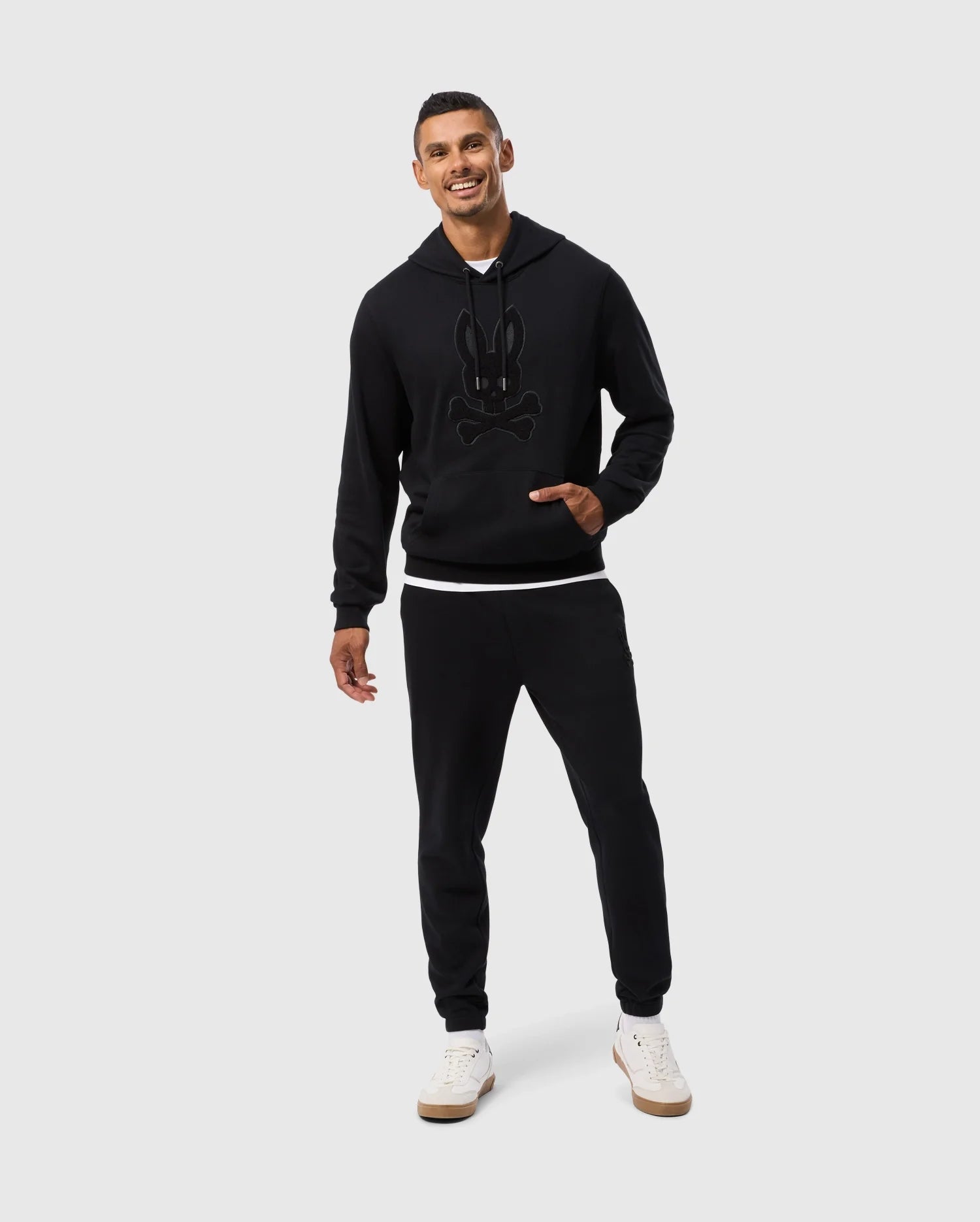 A person smiles, clad in a black MENS PACIFIC CHENILLE HOODIE by Psycho Bunny, featuring subtle chenille embroidery and matching sweatpants, standing against a plain background. They complete the ensemble with white sneakers.
