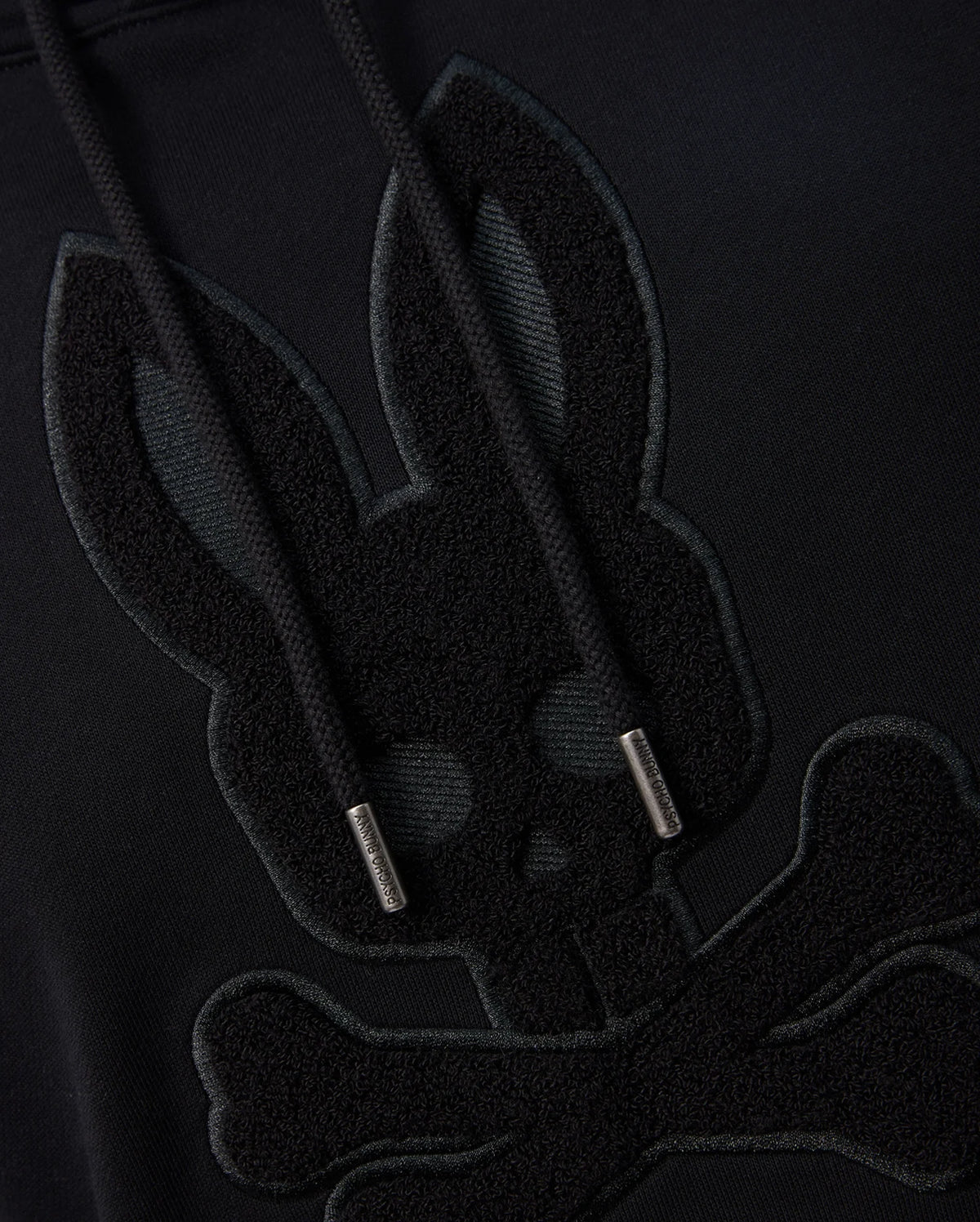 Close-up of the Psycho Bunny MENS PACIFIC CHENILLE HOODIE - B6H930D200 in black, highlighting chenille embroidery featuring a stylized skull with bunny ears atop crossed bones. The cotton knit strings are visible and feature metal-capped tips.