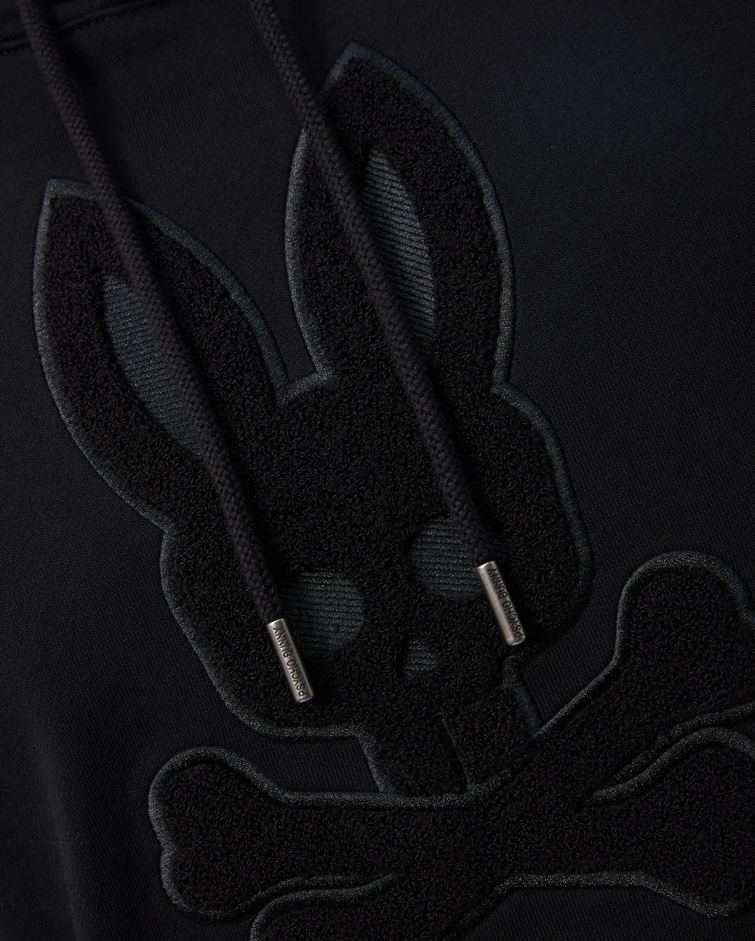 Close-up of the Psycho Bunny MENS PACIFIC CHENILLE HOODIE - B6H930D200 in black, highlighting chenille embroidery featuring a stylized skull with bunny ears atop crossed bones. The cotton knit strings are visible and feature metal-capped tips.