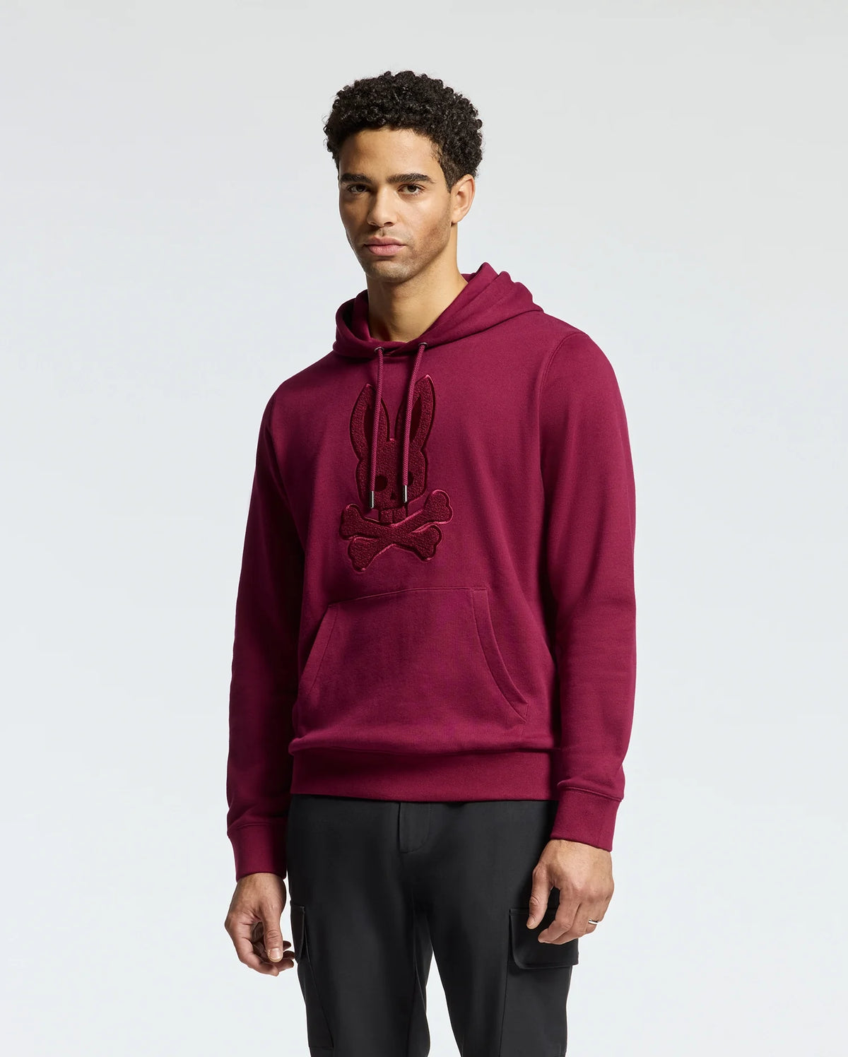 A person dressed in an elegant Psycho Bunny MENS PACIFIC HIGH PILE CHENILLE HOODIE - B6H930D200, adorned with a maroon rabbit and crossbones design, stands against a plain background. The hoodie includes a front pocket and drawstrings for enhanced style and comfort.