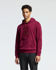 A person dressed in an elegant Psycho Bunny MENS PACIFIC HIGH PILE CHENILLE HOODIE - B6H930D200, adorned with a maroon rabbit and crossbones design, stands against a plain background. The hoodie includes a front pocket and drawstrings for enhanced style and comfort.