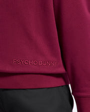 Close-up of a maroon luxurious cotton hoodie, branded as the "MENS PACIFIC HIGH PILE CHENILLE HOODIE - B6H930D200" by Psycho Bunny, with their name embroidered near the hem. It is paired with black pants.