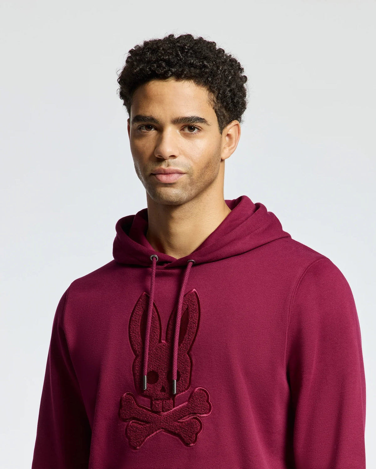 A person wearing a maroon Psycho Bunny MENS PACIFIC HIGH PILE CHENILLE HOODIE - B6H930D200, adorned with a bunny and crossbones design, stands against a plain background. They have short, curly hair and a neutral expression.