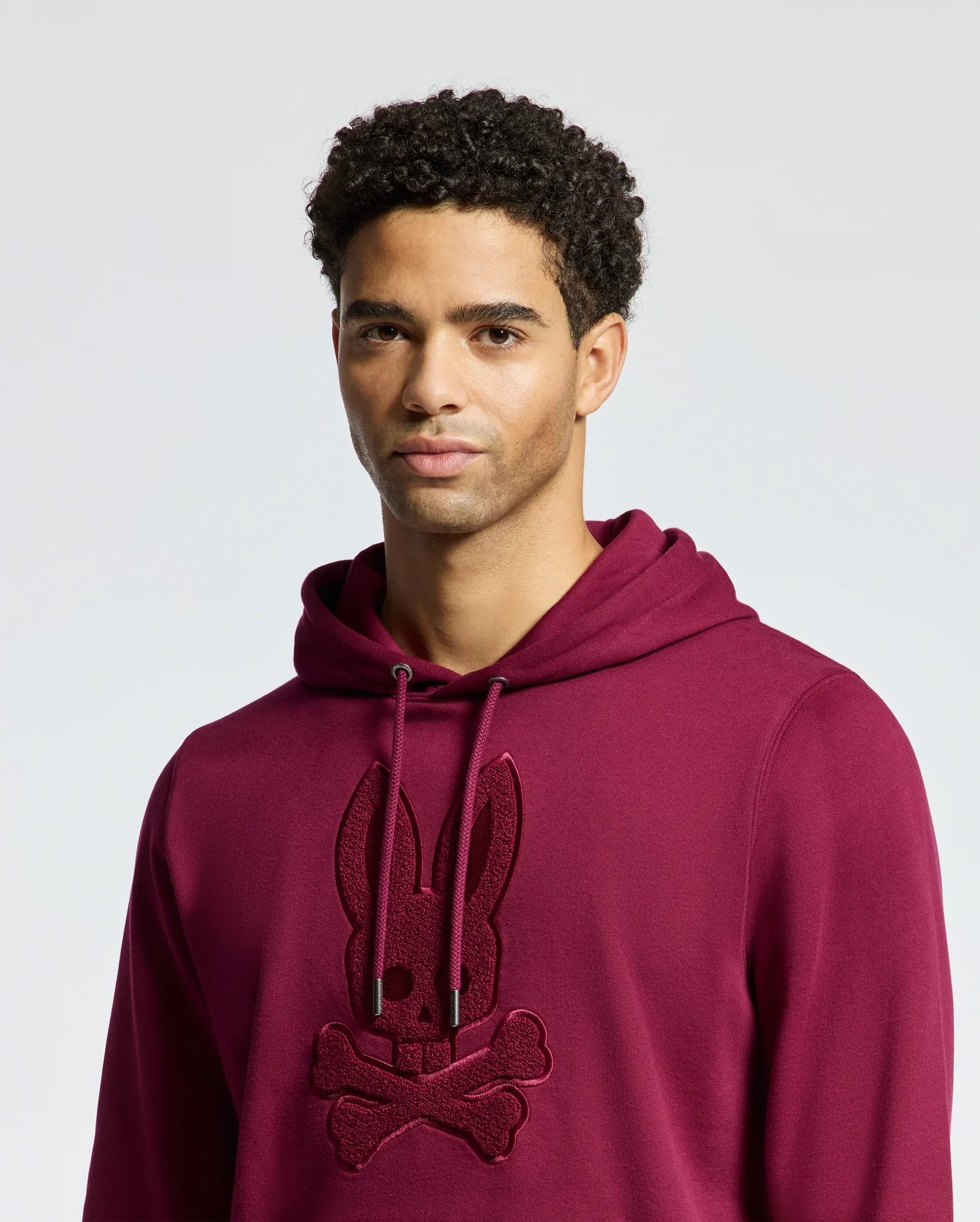 A person wearing a maroon Psycho Bunny MENS PACIFIC HIGH PILE CHENILLE HOODIE - B6H930D200, adorned with a bunny and crossbones design, stands against a plain background. They have short, curly hair and a neutral expression.
