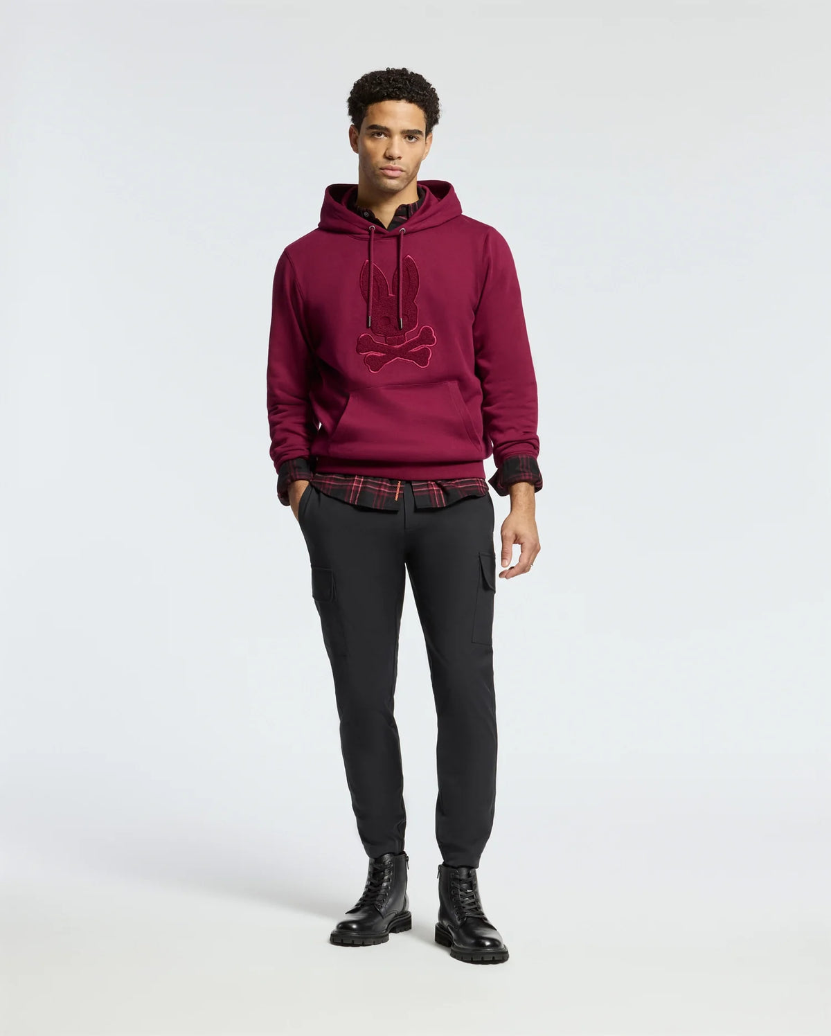 A person stands against a plain background wearing the luxurious MENS PACIFIC HIGH PILE CHENILLE HOODIE by Psycho Bunny, layered over a red plaid shirt. They pair it with black fitted pants and black boots, their hands relaxed by their sides.