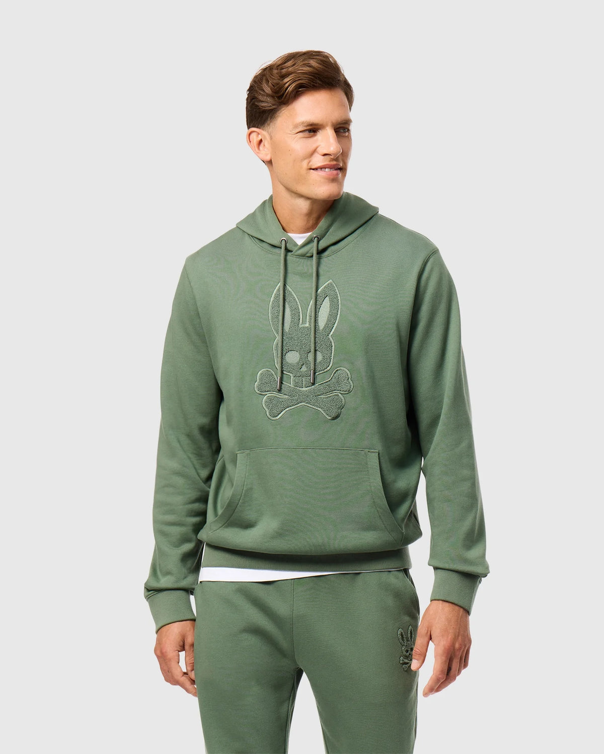 A person wearing the Psycho Bunny Men's Pacific Chenille Hoodie - B6H930D200, featuring a bunny and crossbones design, stands smiling. The outfit is crafted from French terry and includes matching green sweatpants against a light gray background.