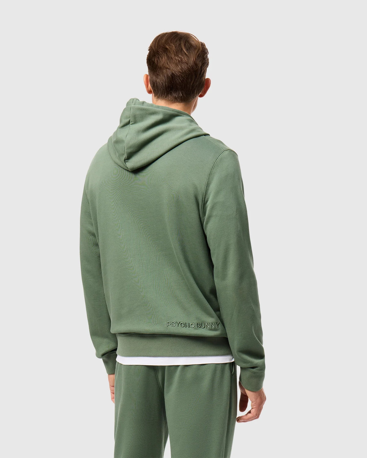 A person wearing a green Psycho Bunny hoodie, specifically the Men's Pacific Chenille Hoodie (B6H930D200), paired with matching sweatpants is seen from behind. The French terry hoodie showcases "Psycho Bunny" in chenille embroidery on the lower back. The individual, who has short brown hair, stands against a light gray background.
