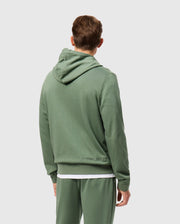 A person wearing a green Psycho Bunny hoodie, specifically the Men's Pacific Chenille Hoodie (B6H930D200), paired with matching sweatpants is seen from behind. The French terry hoodie showcases "Psycho Bunny" in chenille embroidery on the lower back. The individual, who has short brown hair, stands against a light gray background.