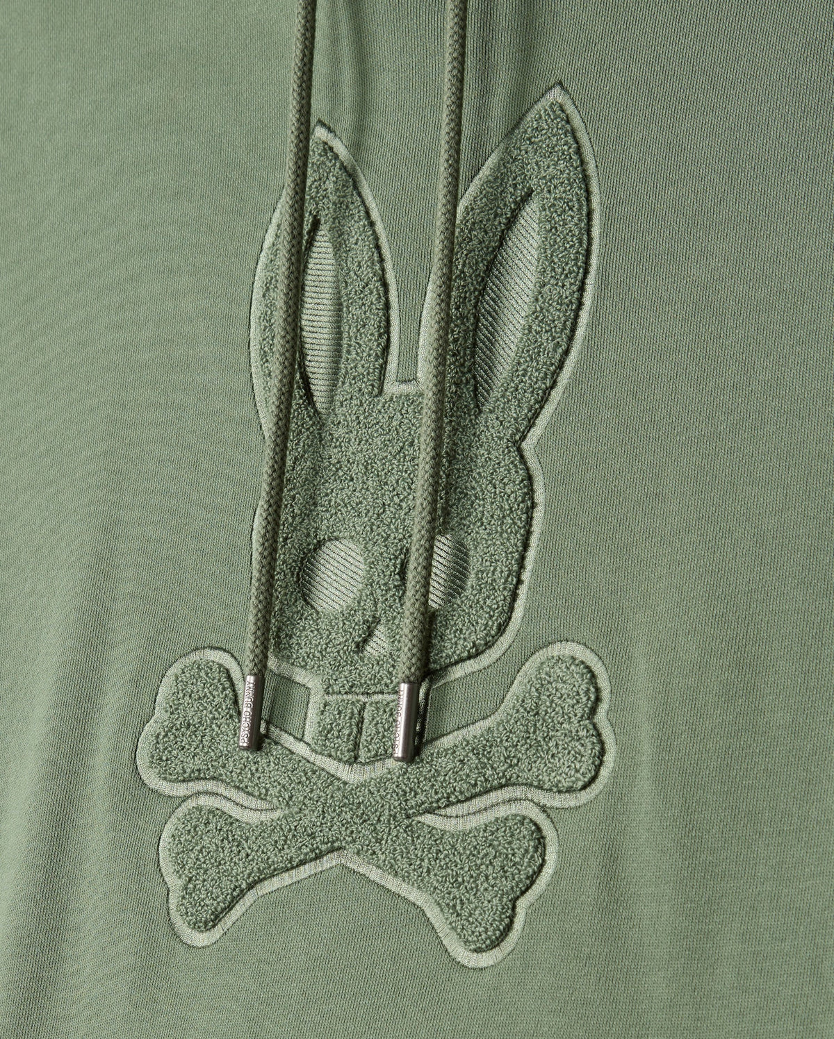 The MENS PACIFIC CHENILLE HOODIE - B6H930D200 by Psycho Bunny features green fabric embellished with chenille embroidery of a stylized bunny skull and crossbones. Cord drawstrings gracefully drape over the design, adding an elegant touch.