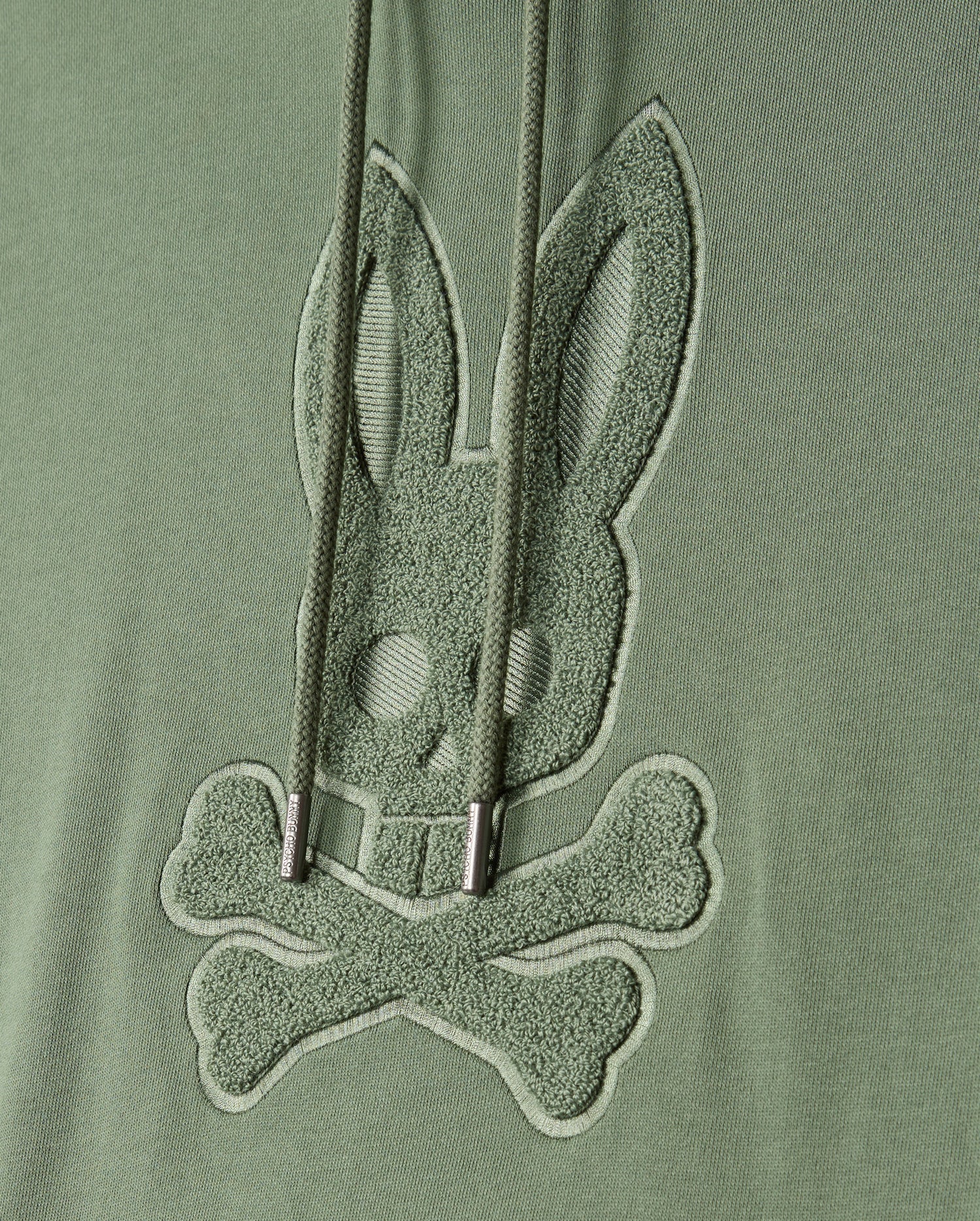 The MENS PACIFIC CHENILLE HOODIE - B6H930D200 by Psycho Bunny features green fabric embellished with chenille embroidery of a stylized bunny skull and crossbones. Cord drawstrings gracefully drape over the design, adding an elegant touch.